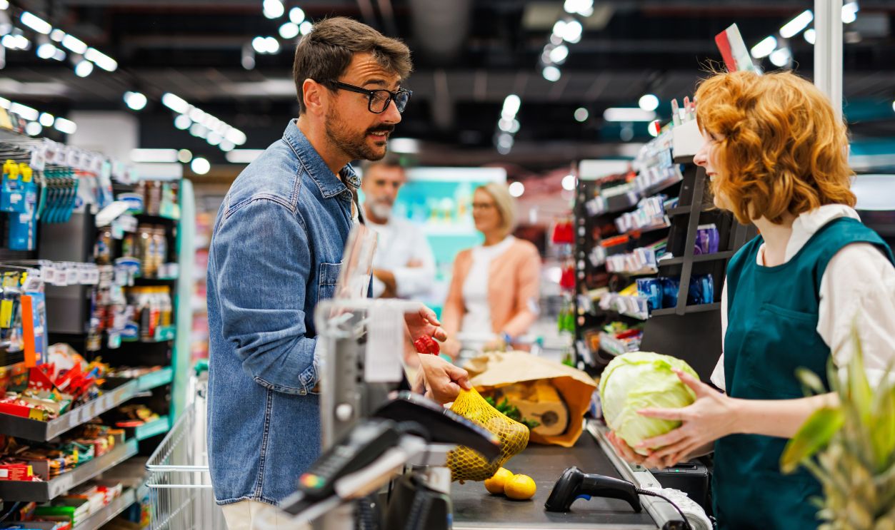 Shutterstock-2546757641, Supermarket cashier scanning groceries at the checkout counter while talking with a male customer, other customers waiting in line behind