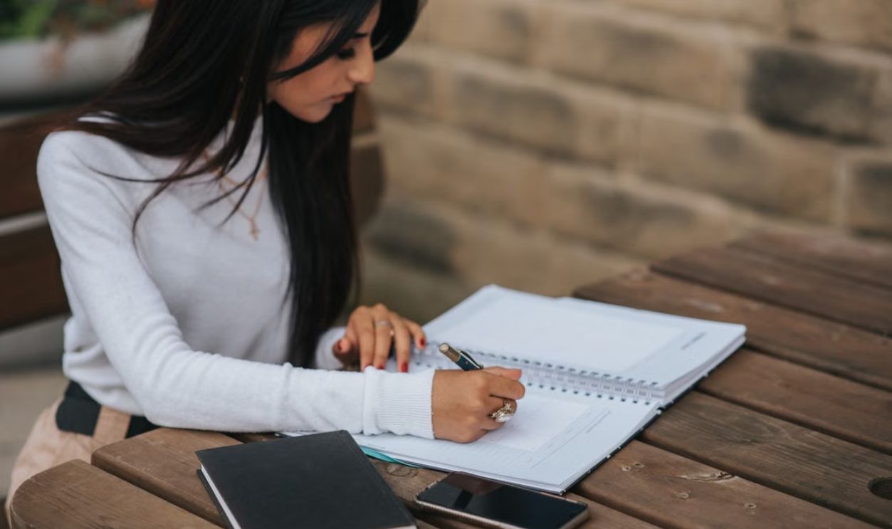Focused young woman writing in a notebook at an outdoor table, with a smartphone nearby