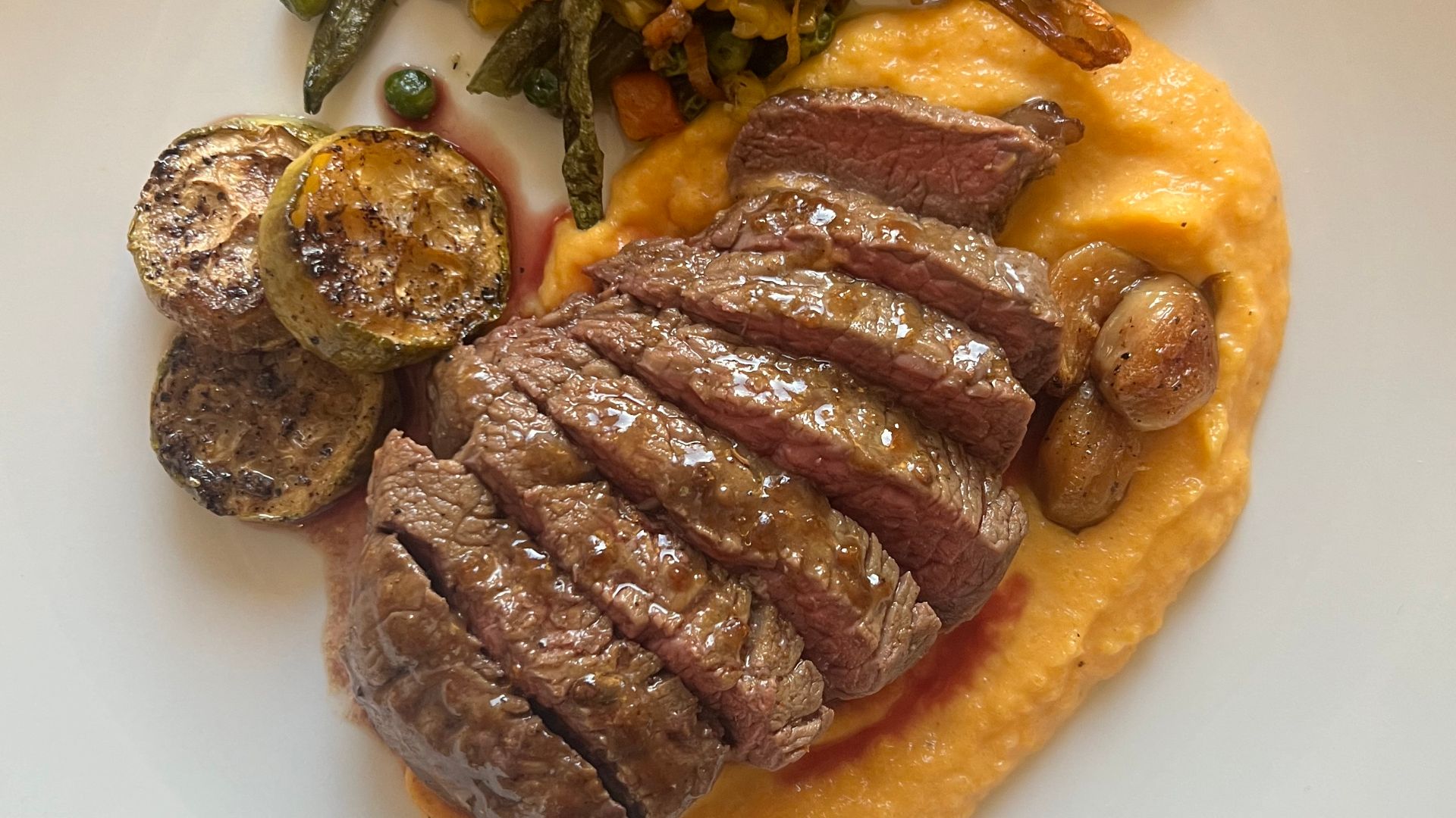 High-quality photograph by Liat Portal showing a homemade meal of sliced steak cooked on a steel skillet, served over mashed butternut squash and sweet potatoes, alongside roasted grey zucchini, grilled garlic, and mixed vegetables. This image documents a