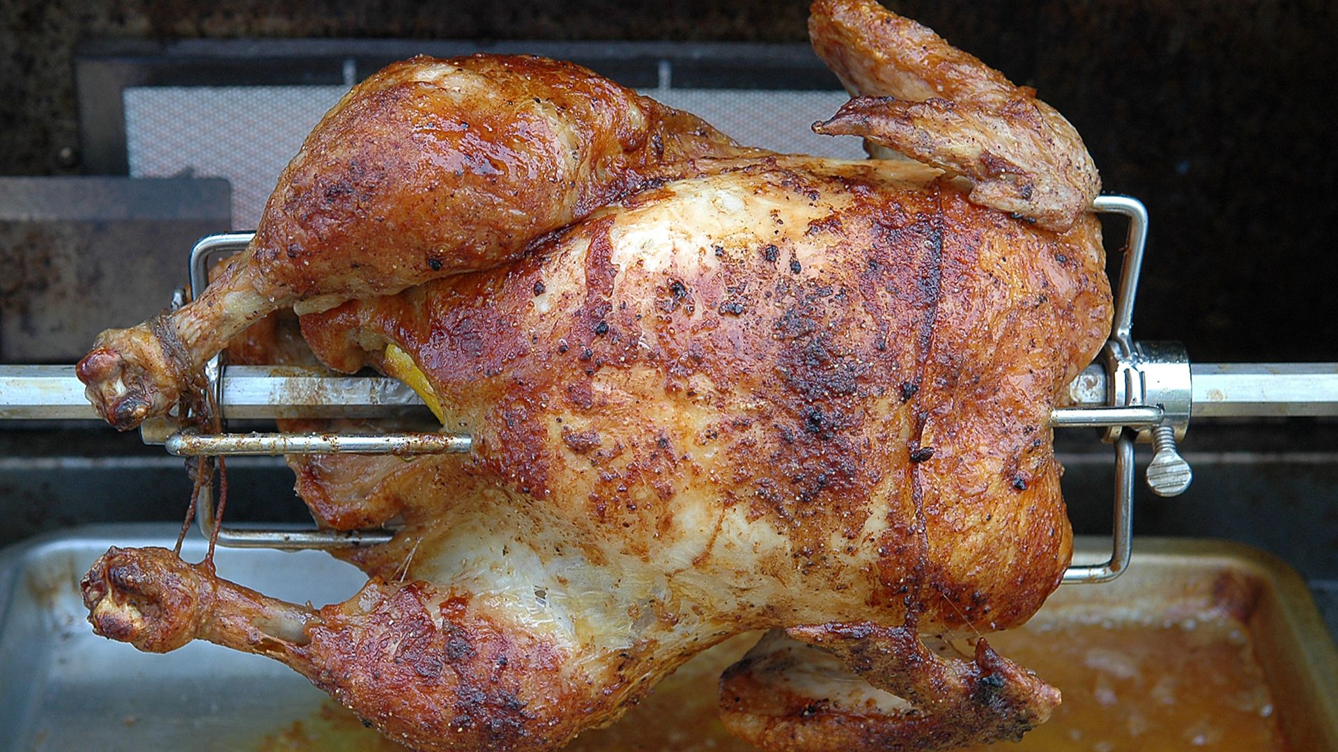 Chicken cooking on a gas grill rotisserie. The yellow protruding from the cavity is a pierced whole lemon.