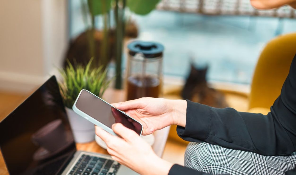 Woman working remotely from home, using a smartphone and laptop at a cozy desk with a plant and cat in background
