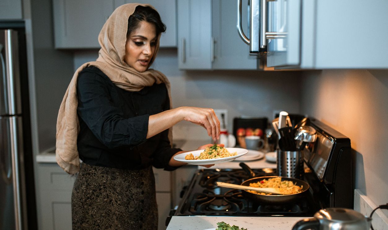 A woman in a hijab garnishes food in a modern kitchen setting, adding coriander