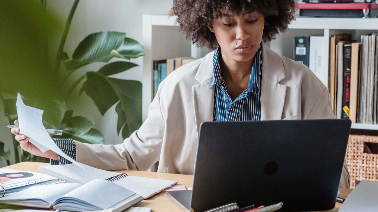 Woman working at a home office desk with a laptop, plants, and paperwork around