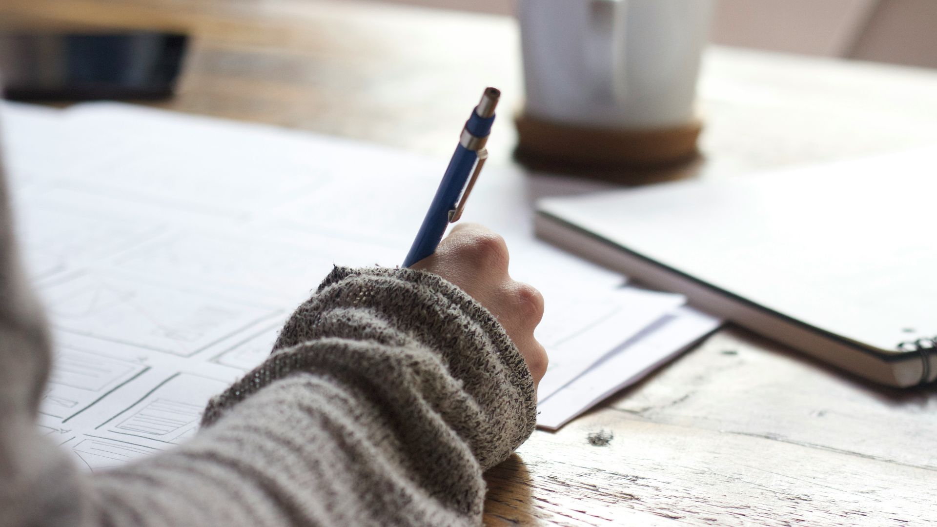 person writing on brown wooden table near white ceramic mug