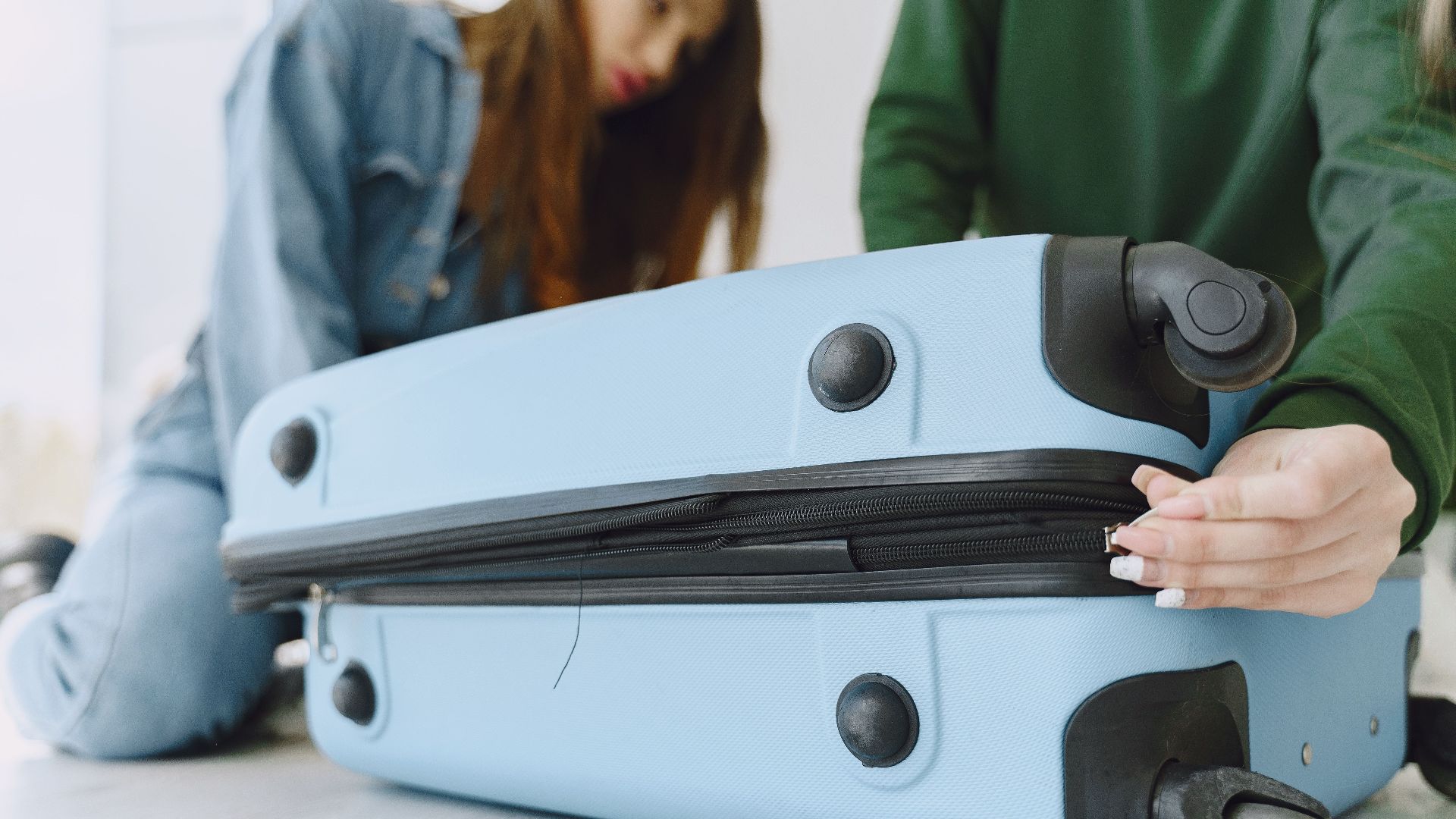 From below of crop concentrated young woman in denim outfit closing suitcase sitting on floor with anonymous friend while preparing for trip