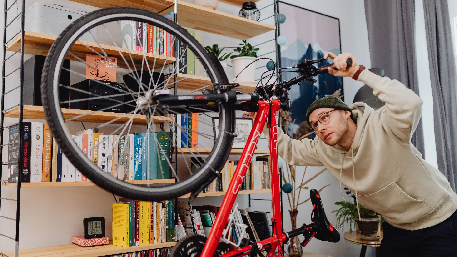 A man in a hoodie adjusts a bicycle indoors in front of a bookshelf.