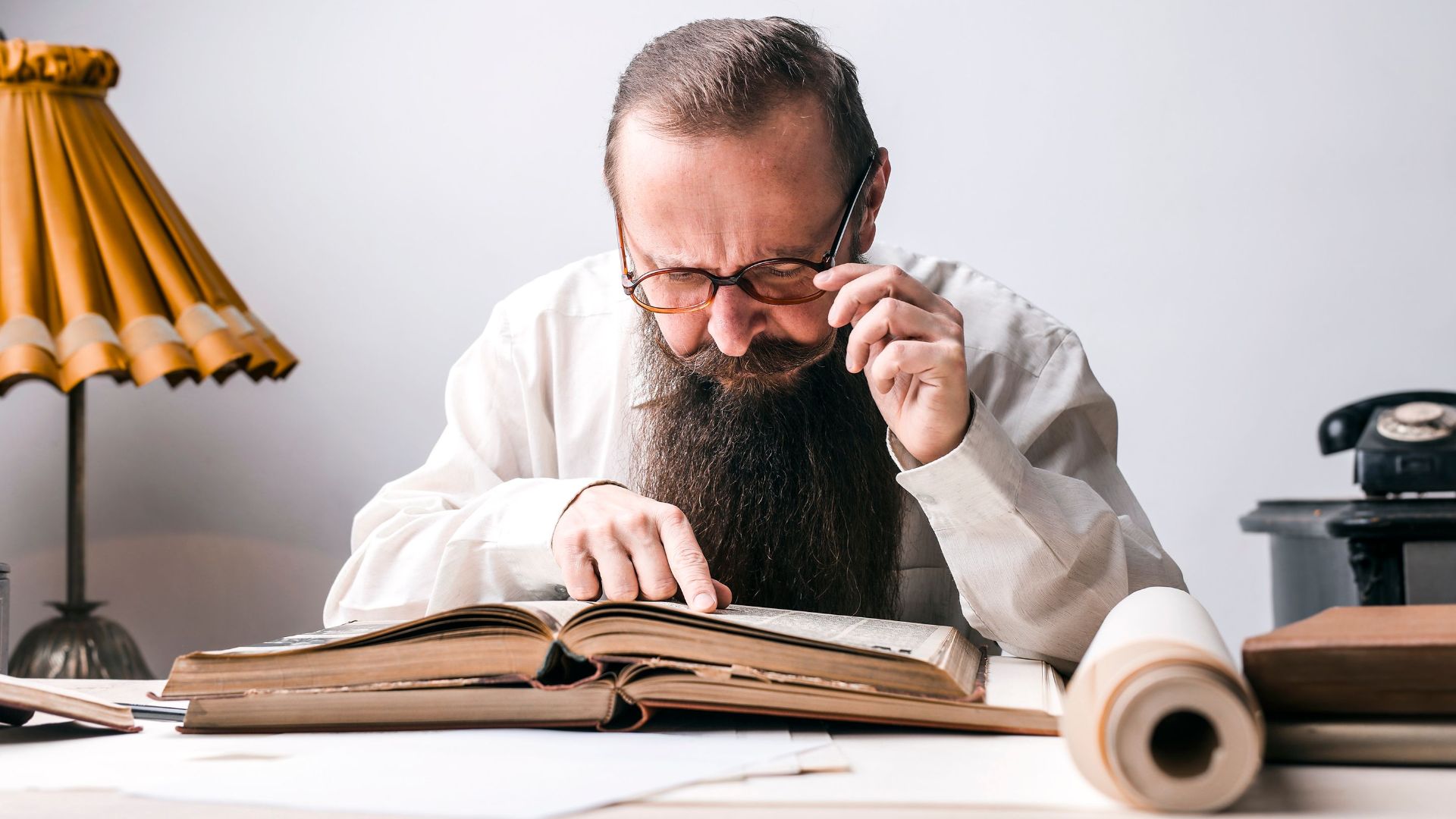 Bearded man with eyeglasses focused on reading a large book at a vintage desk.