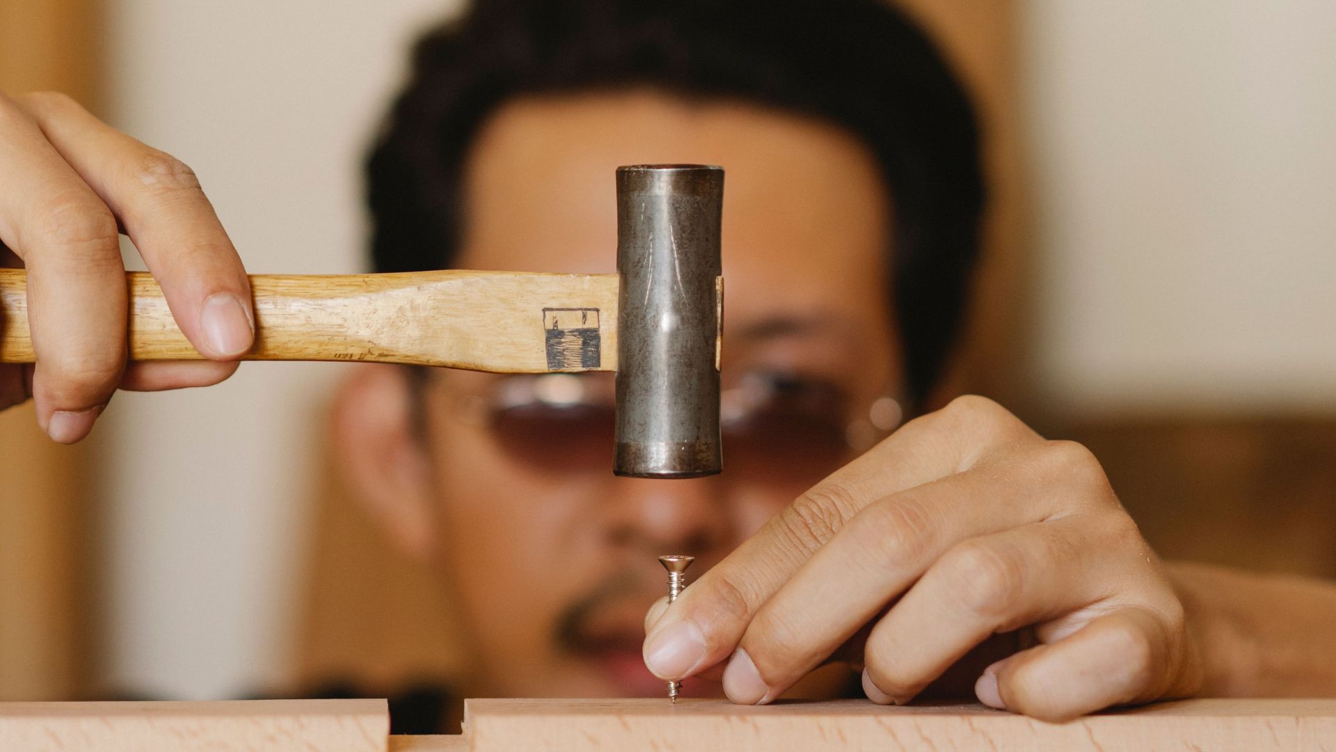 Young ethnic serious male in sunglasses hammering nail in wooden beam in workshop