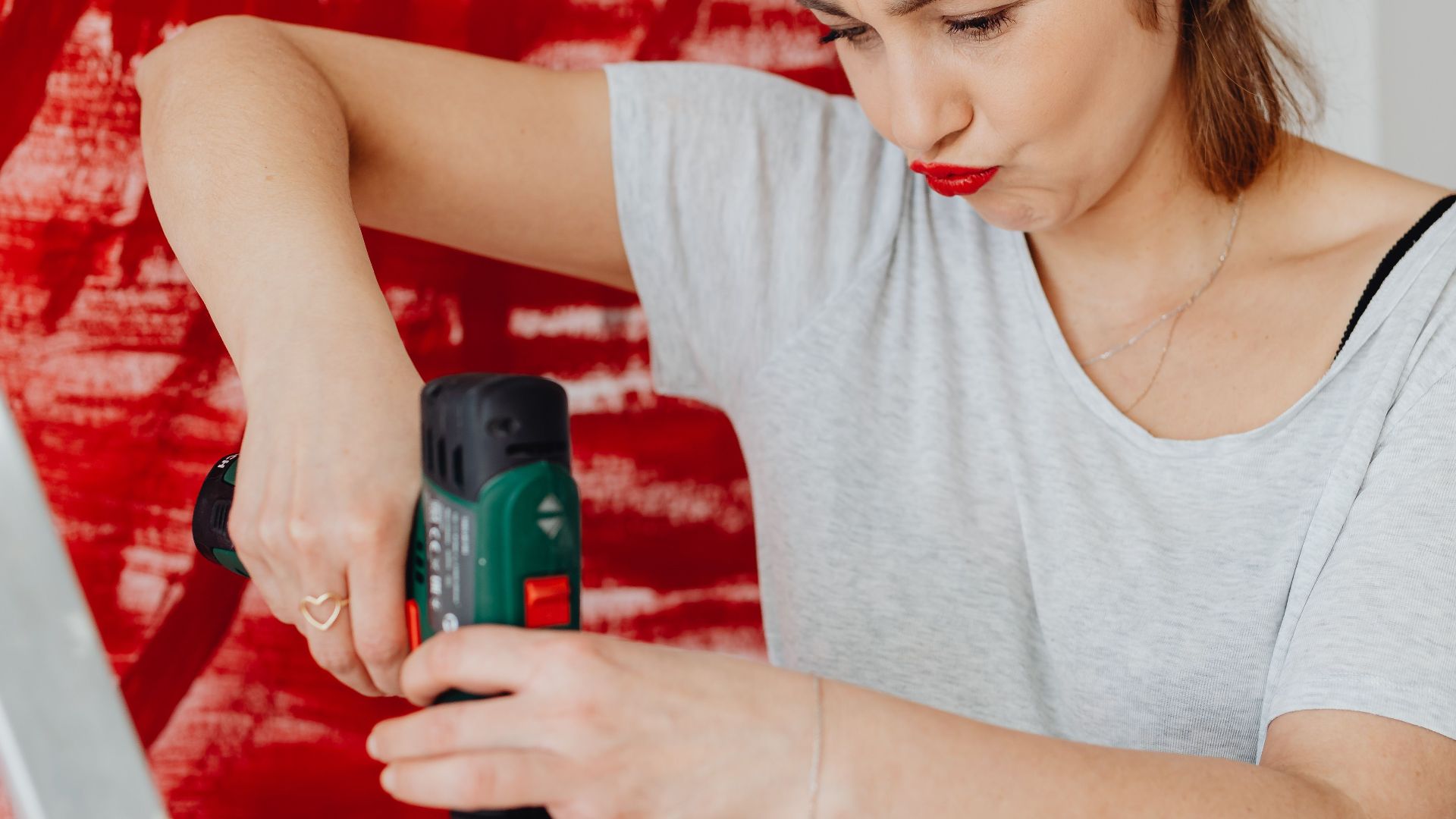 A woman on a ladder uses a drill on an art project with a red abstract background.