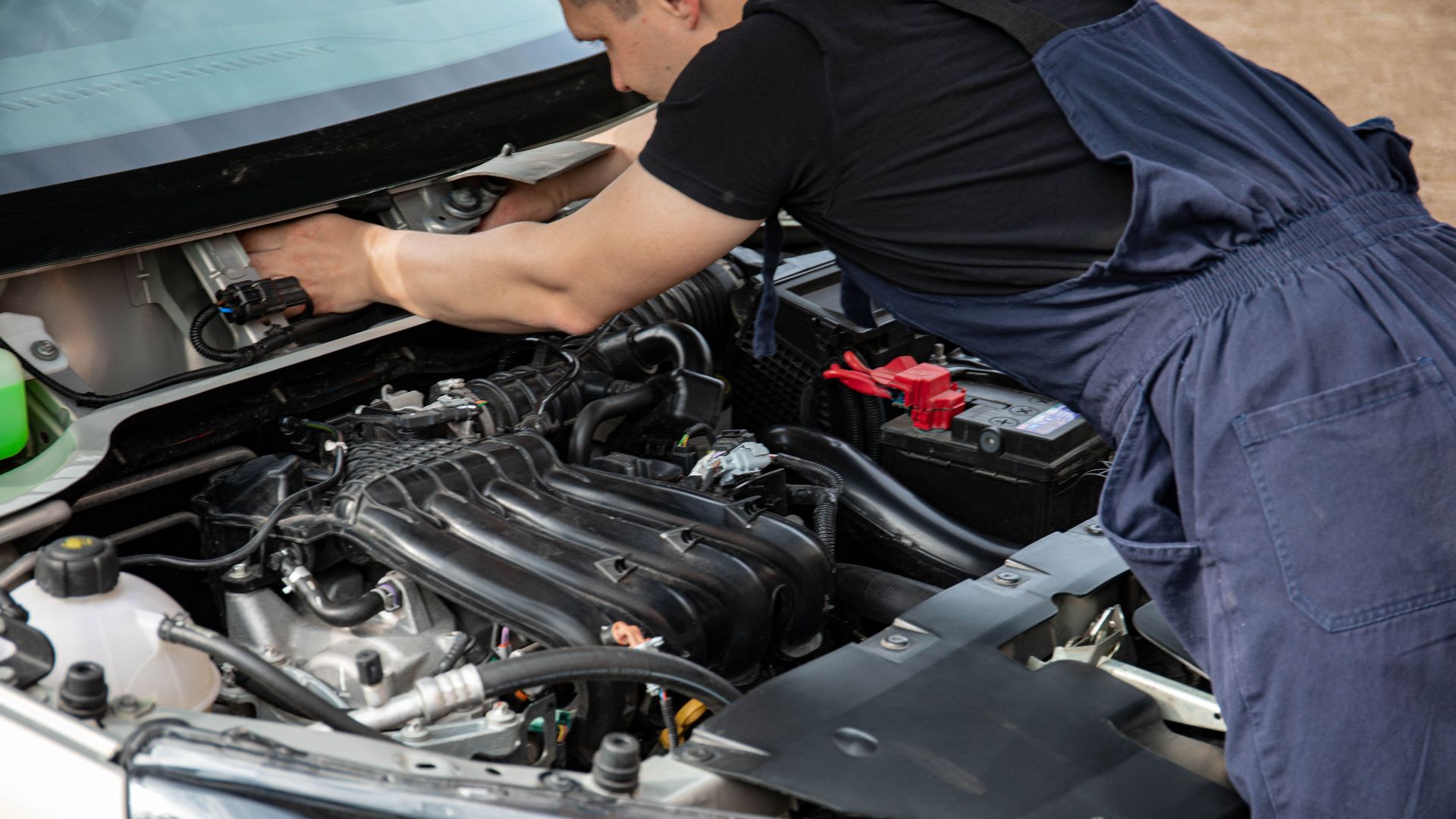 Mechanic in blue overalls working on car engine with open hood outdoors during the day.