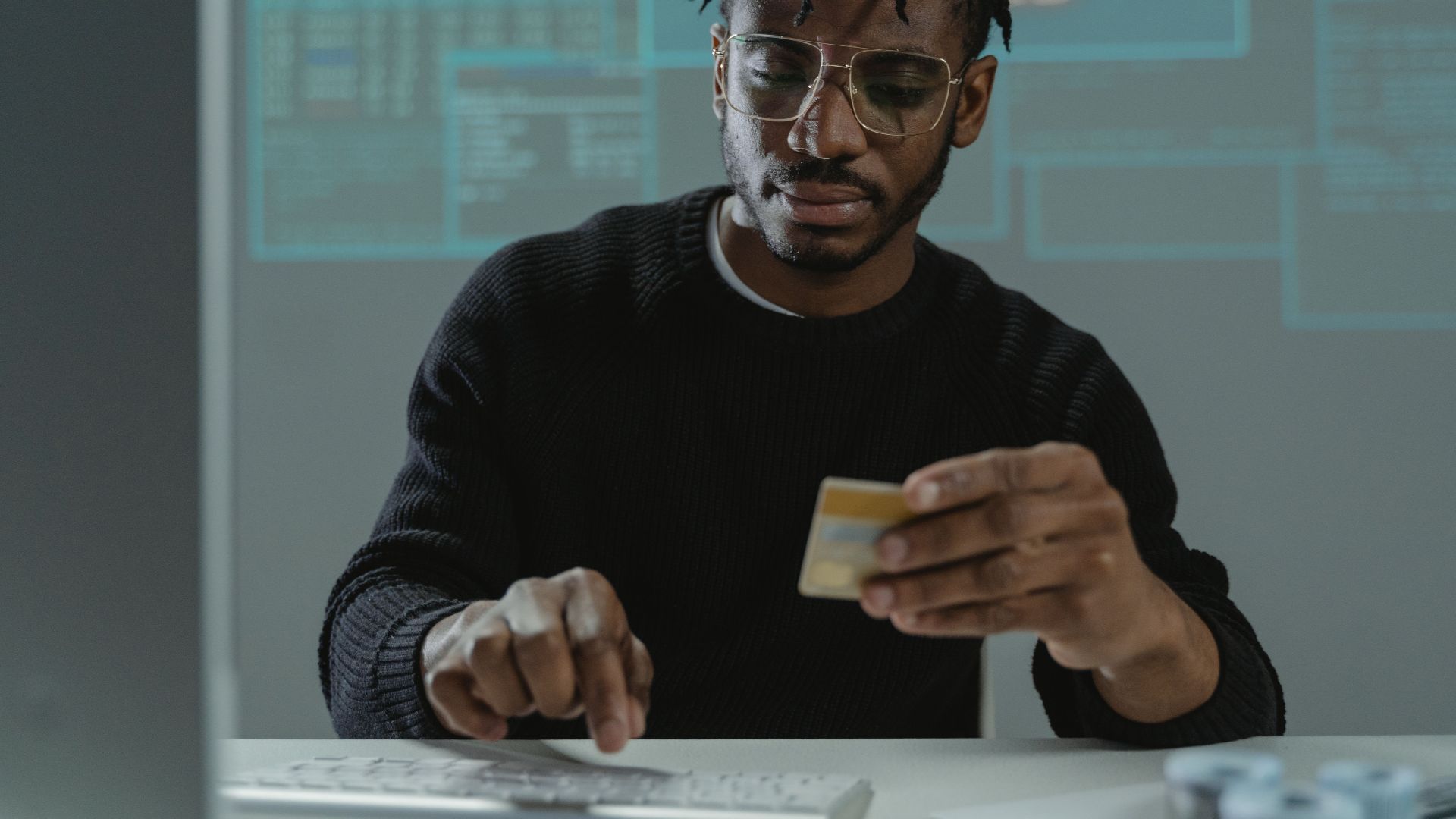 Man with glasses holding a bank card while typing on a keyboard, sitting in front of a computer.