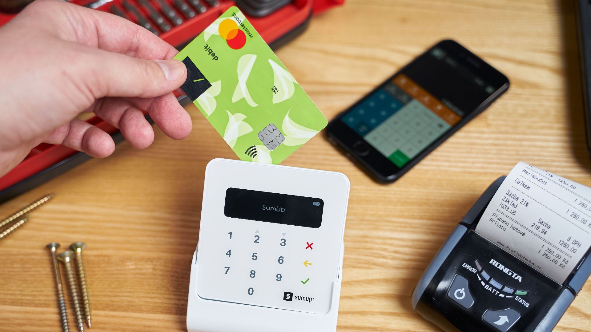Faceless customer paying by small bright plastic card while using cash register among smartphone and tools on wooden table in bright studio