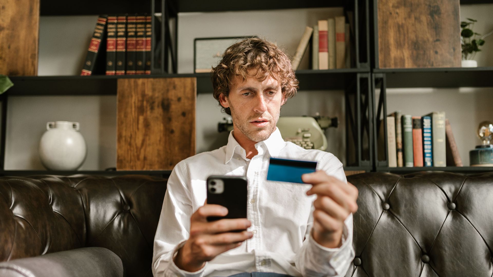 A man sitting on a leather sofa using a smartphone and credit card for online shopping.
