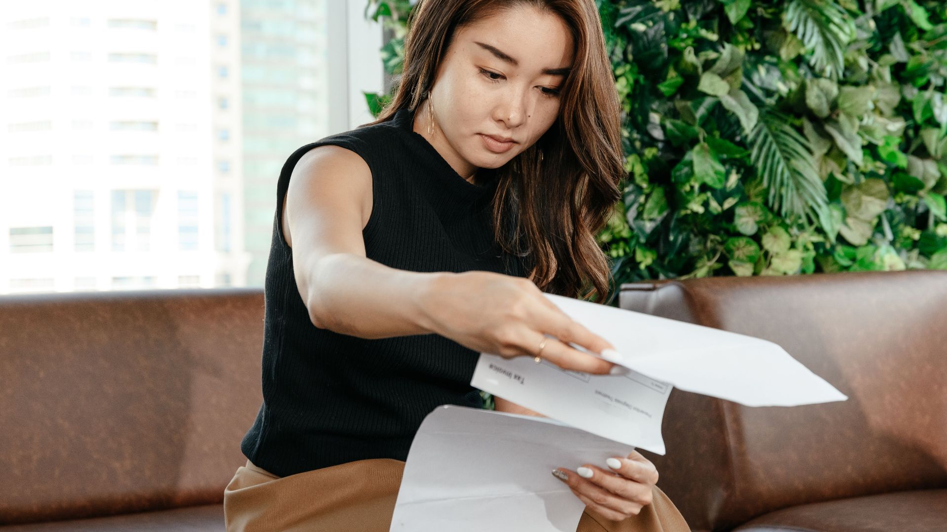 Focused Asian female turning pages of document while sitting on sofa during paperwork in modern workspace with green deciduous plant