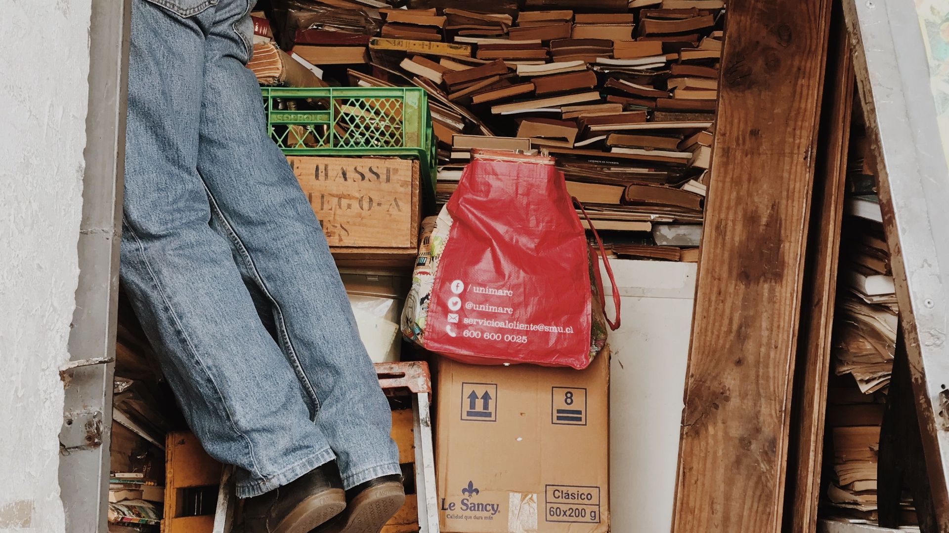 A person climbing a ladder in a cluttered storage area filled with books and boxes.