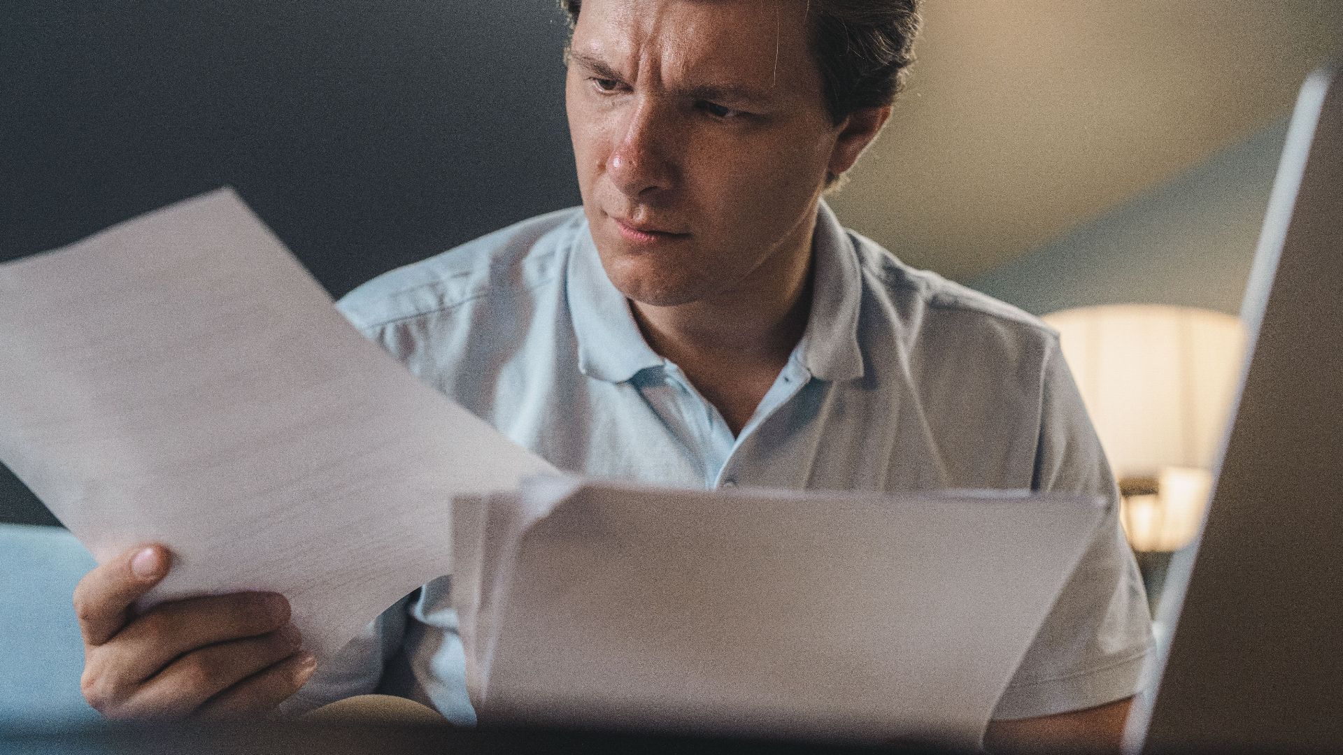 A man intently examines papers, seated indoors under warm lighting, focusing on his work.
