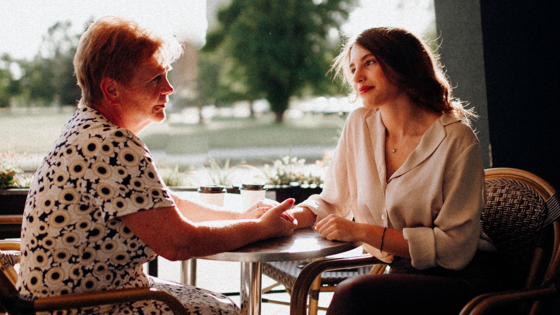 A mother and daughter share a heartfelt moment while sitting at a café table outdoors.