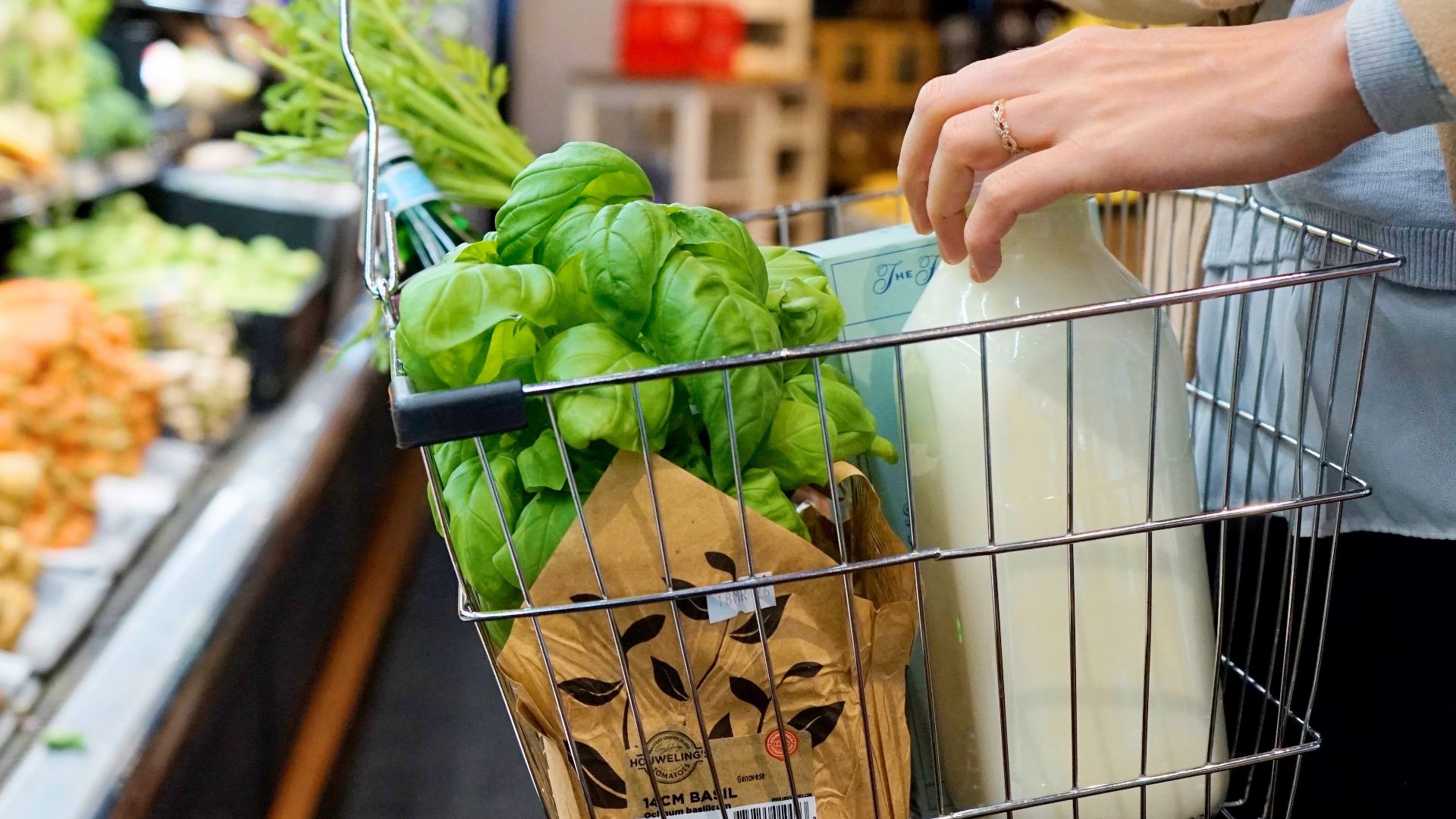 A person shops in a grocery store with a basket containing fresh greens, milk, and herbs.