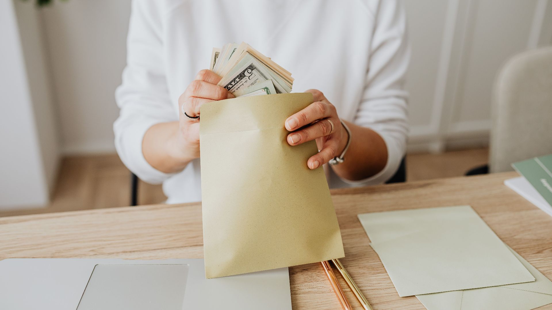 Close-up of person holding envelopes with cash at a wooden desk indoors.