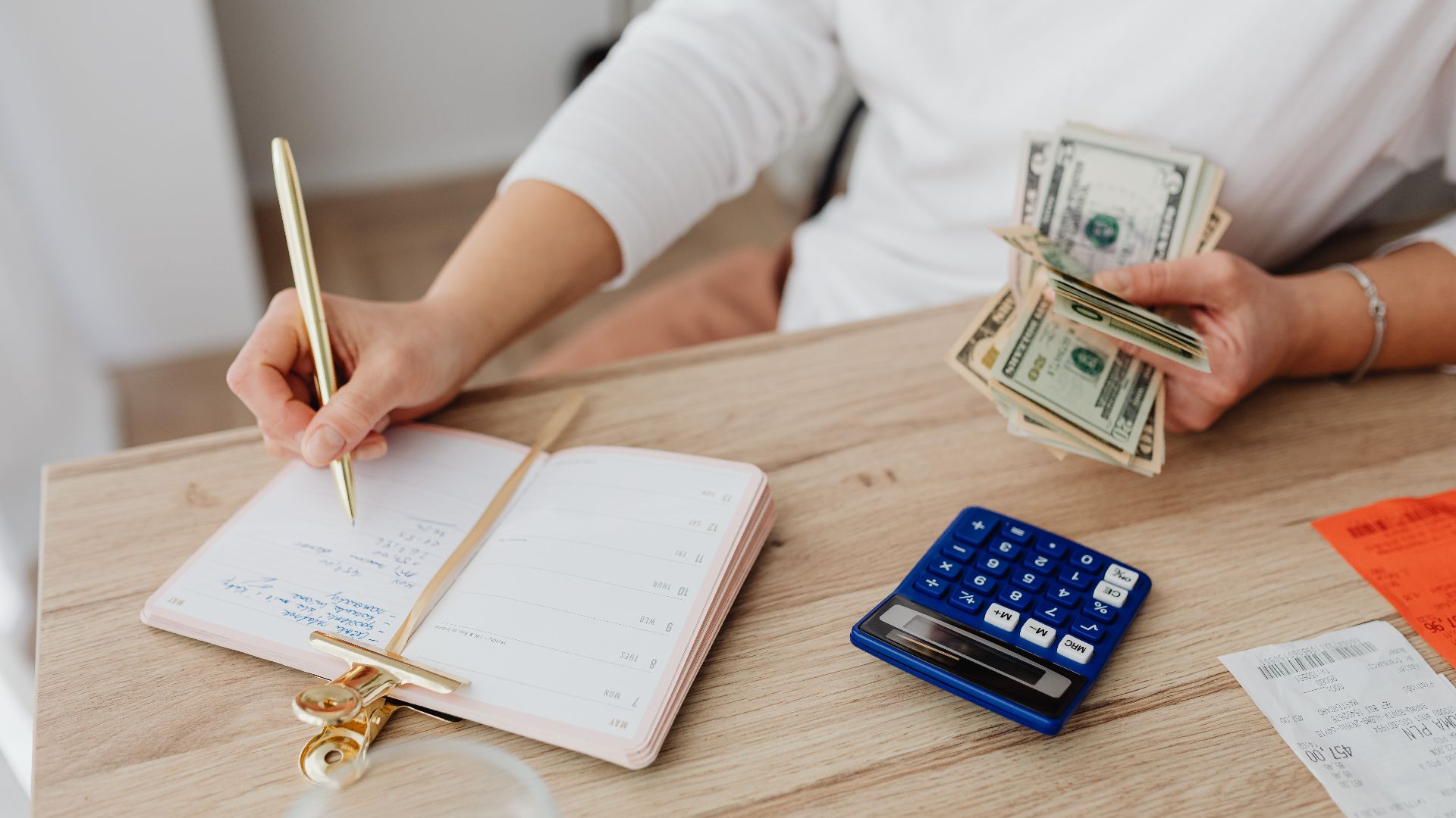 Adult holding cash and writing in planner while using a calculator at home.