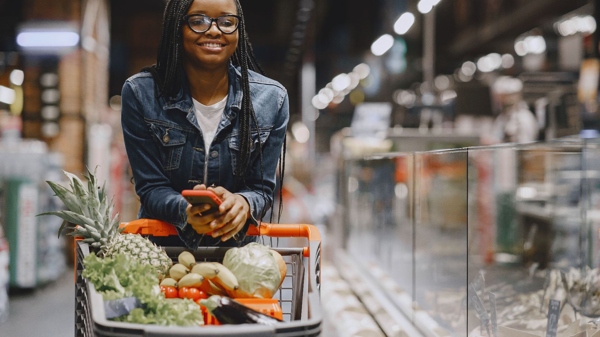 A woman smiles while shopping for fresh produce in a supermarket, using a smartphone.