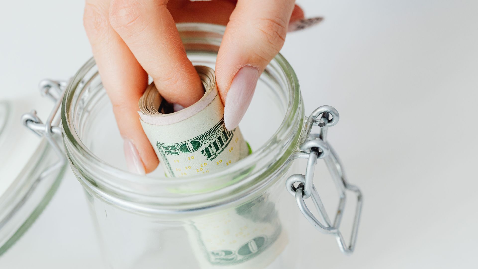 A close-up of a hand placing rolled dollars into a glass jar, symbolizing savings.