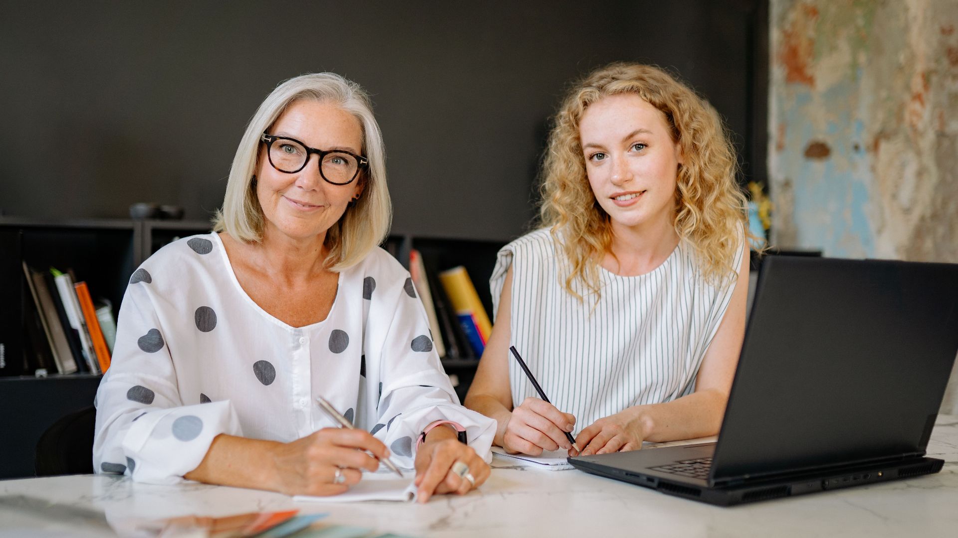 Two women smiling and working together in a modern office setting with a laptop and documents.