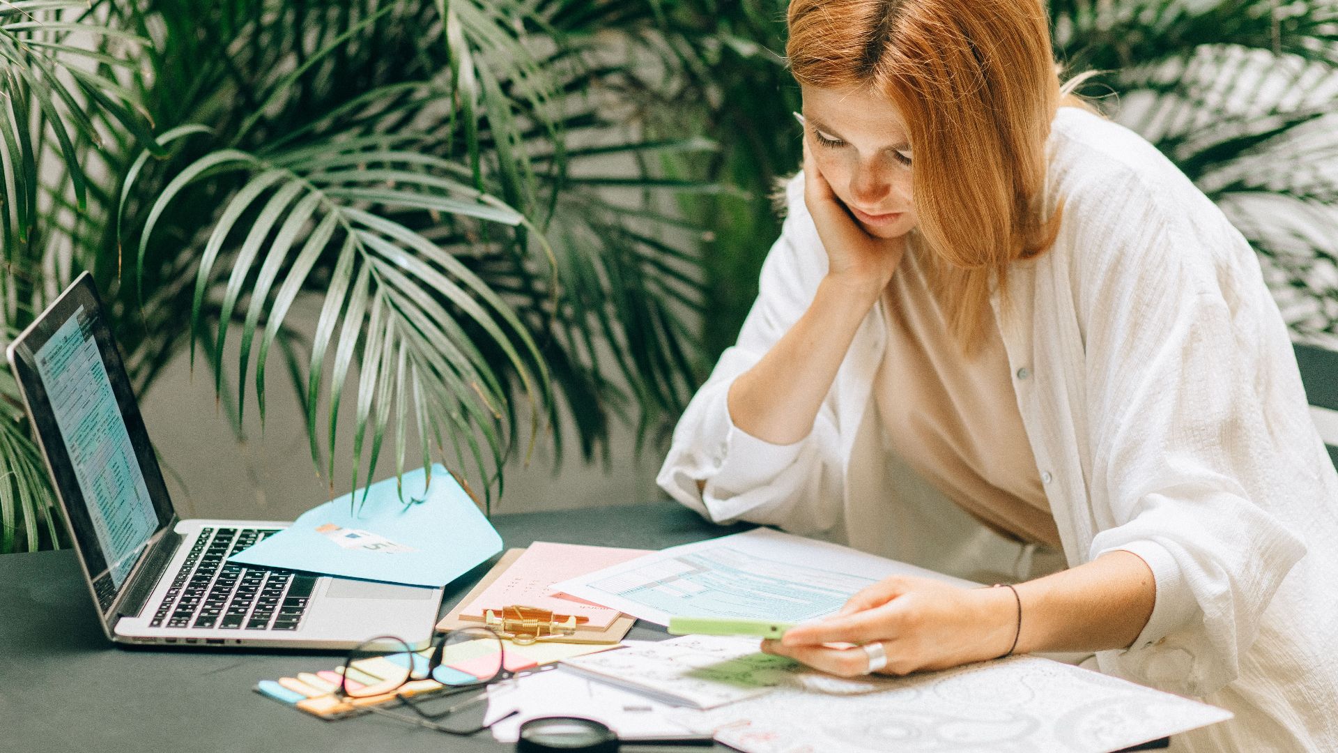 Woman in a thoughtful pose reviewing documents on a table with a laptop, surrounded by greenery.