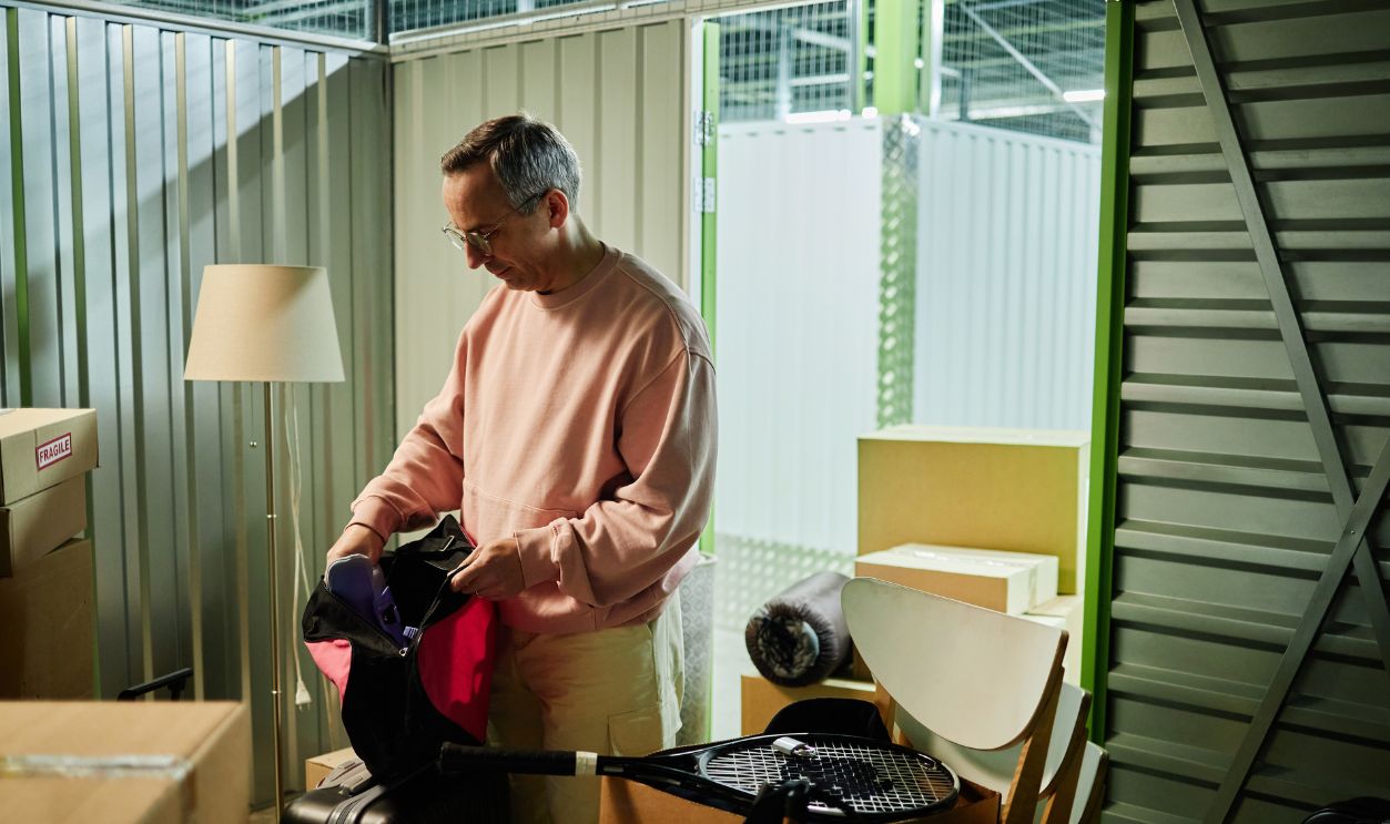 Middle aged Caucasian man sorting belongings in storage unit, holding bag while standing among packed boxes and various household items, focused on organizing personal possessions