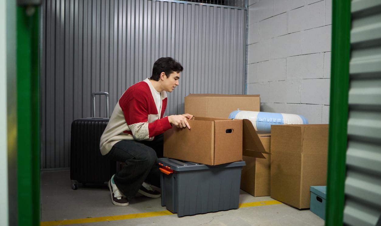 Young adult Caucasian man crouching in storage unit organizing cardboard boxes and plastic containers, focusing on sorting belongings, suitcase standing nearby, industrial setting