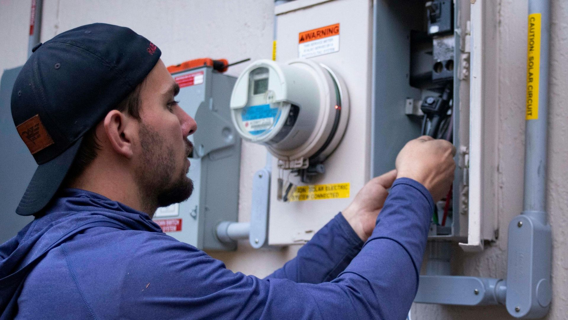 A person in a blue hoodie and baseball cap working on an electrical meter