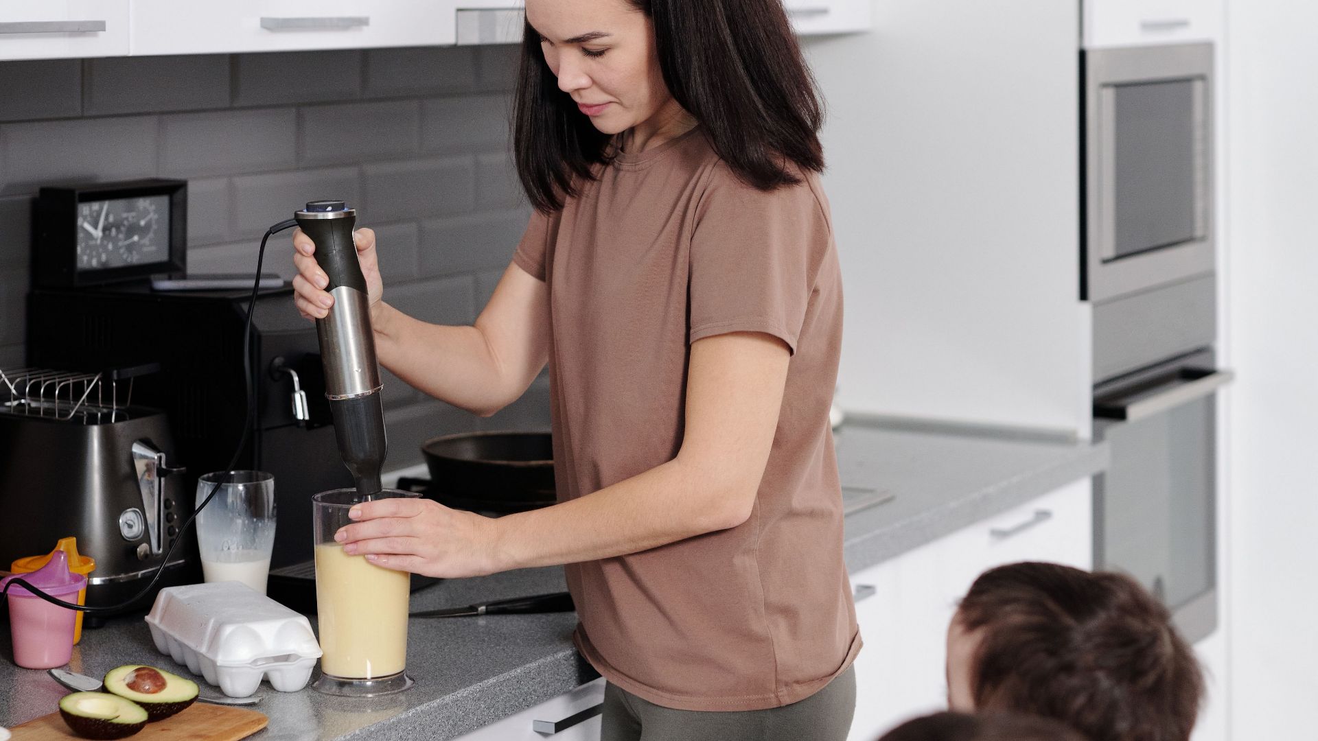 A mother prepares a smoothie with her two young sons in a modern kitchen setting.