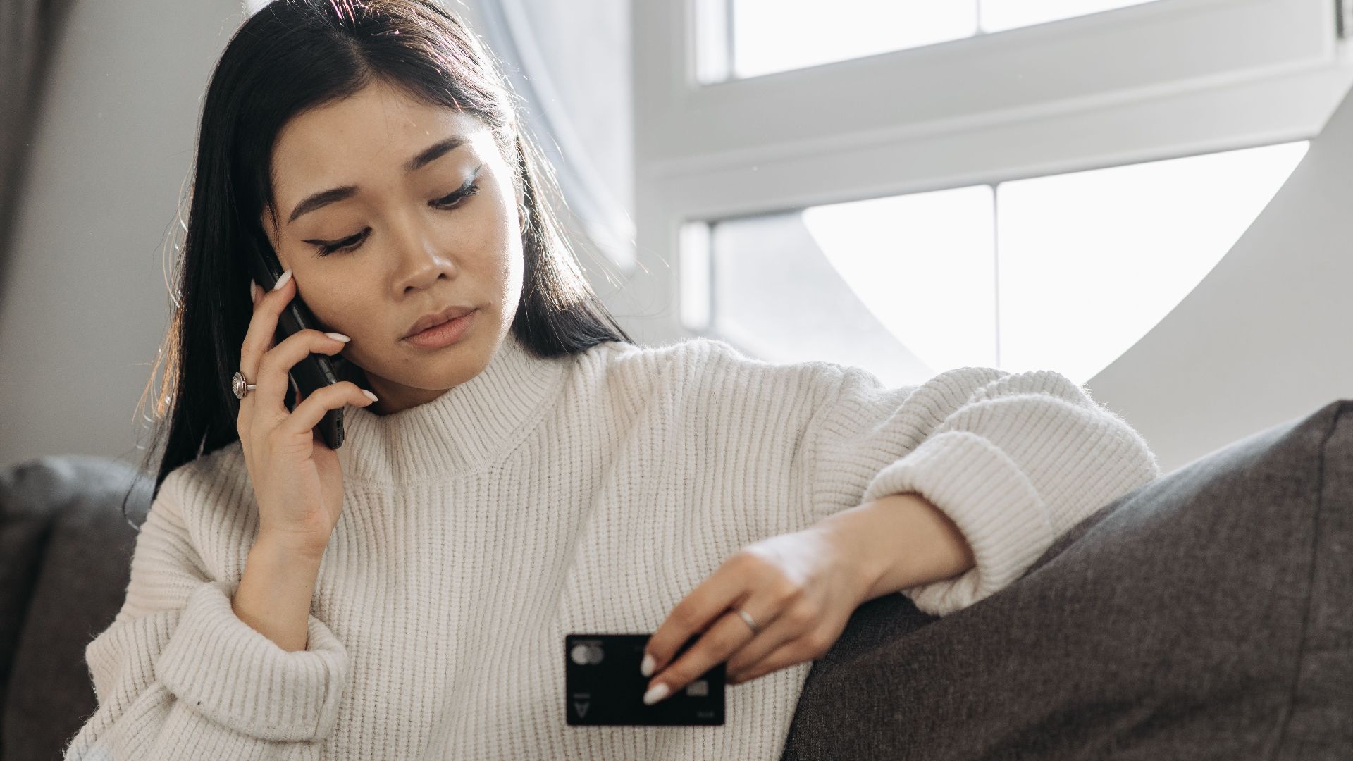 A woman sits on a sofa, multitasking with a phone and credit card, focusing on online shopping.
