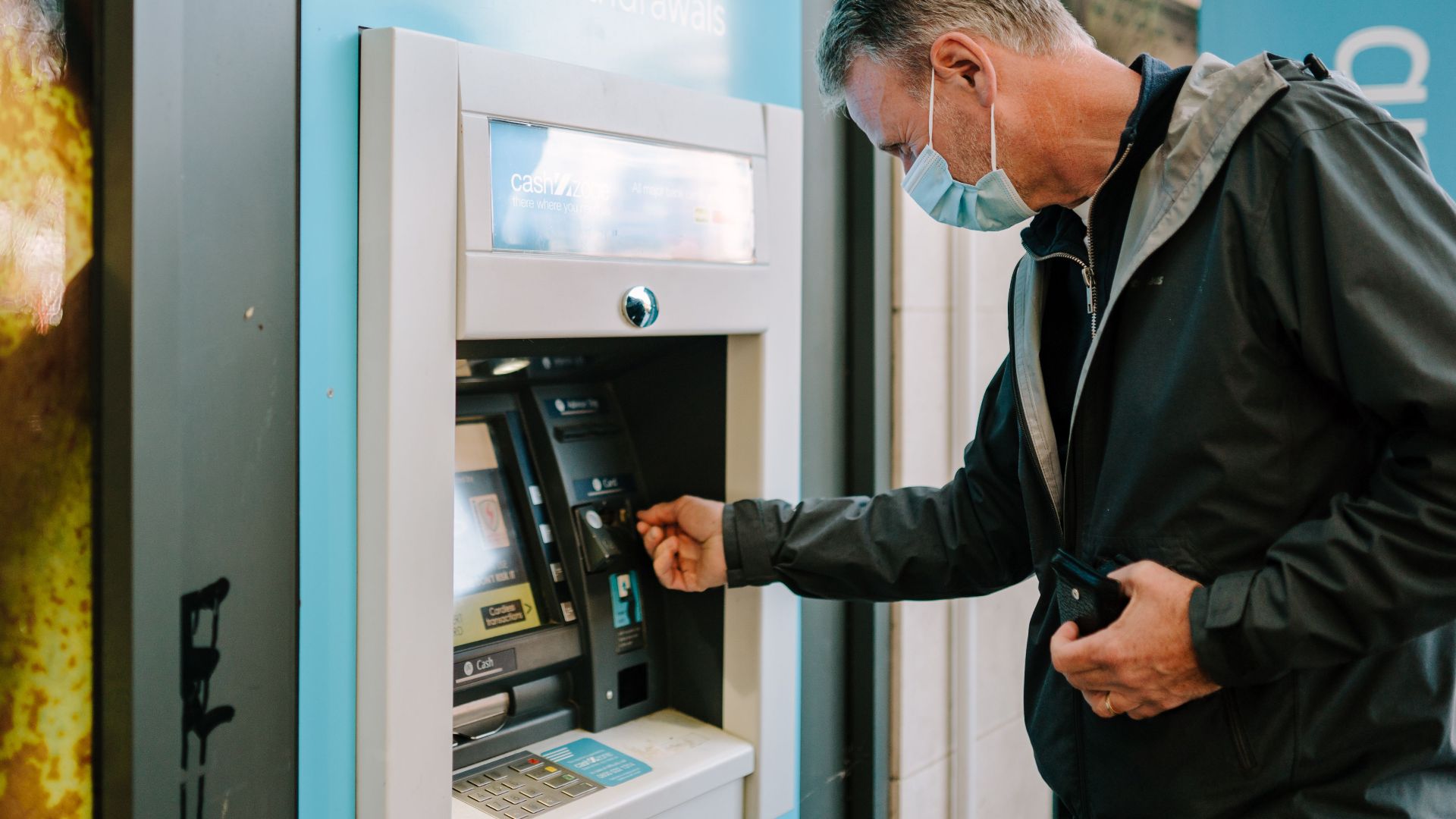 A man wearing a mask using an ATM machine outdoors for cash withdrawal.