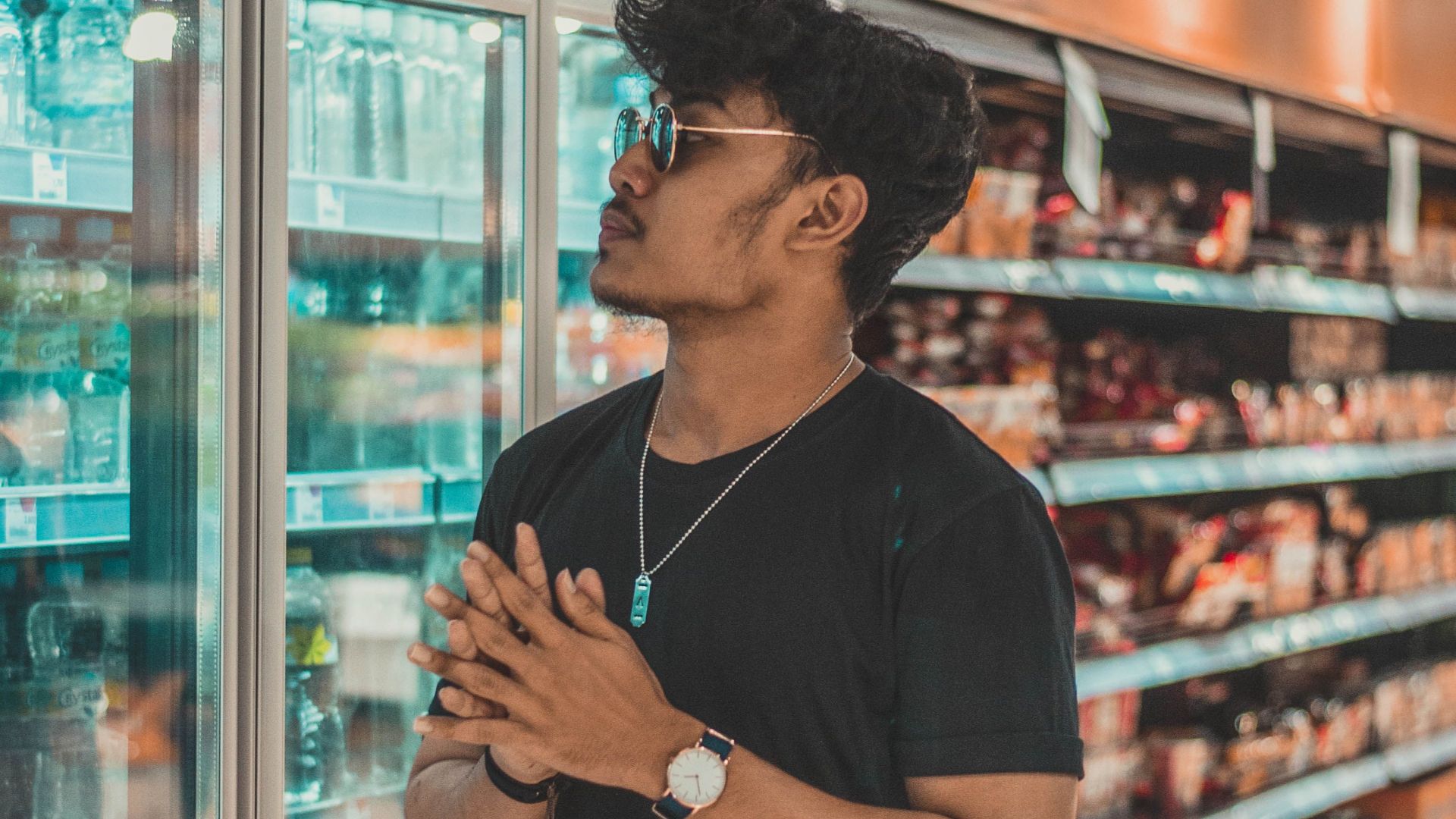 Young calm ethnic male in black shirt and trendy sunglasses standing near refrigerator in modern supermarket and looking at goods
