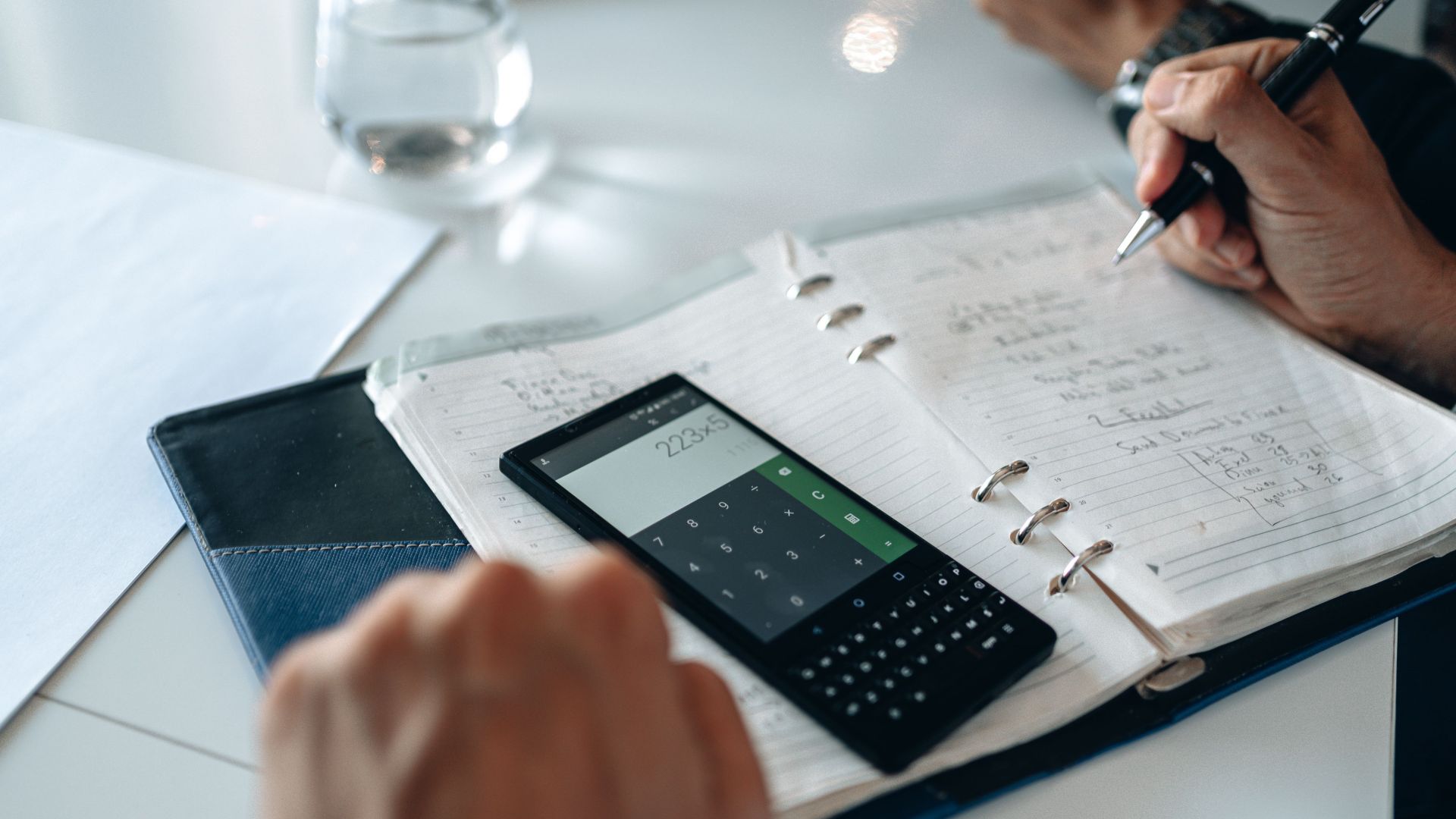 Close-up of hands writing in notebook with smartphone calculator on the table.