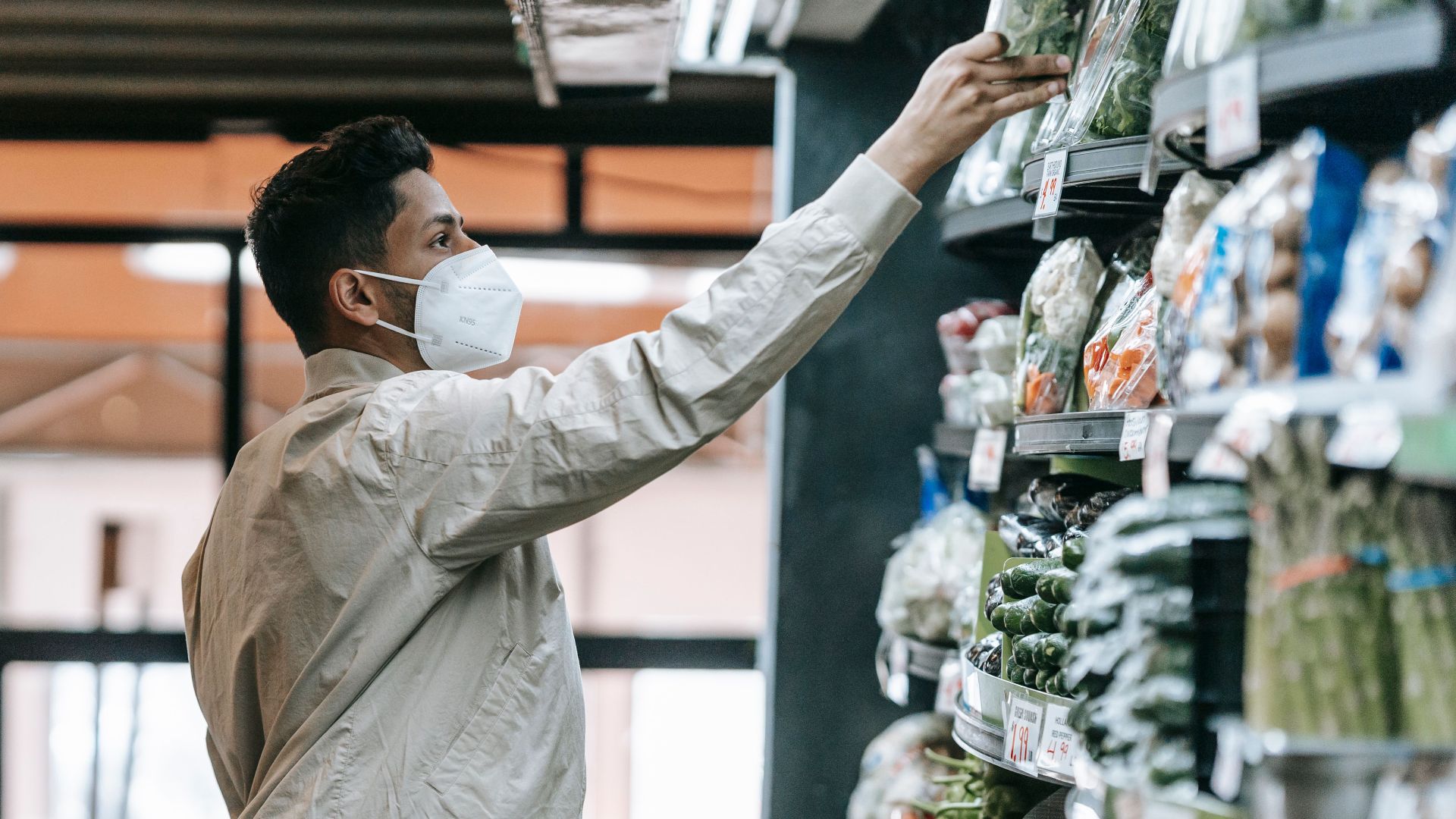 Side view of Indian male customer in protective mask picking fresh products from stall while shopping in supermarket during pandemic