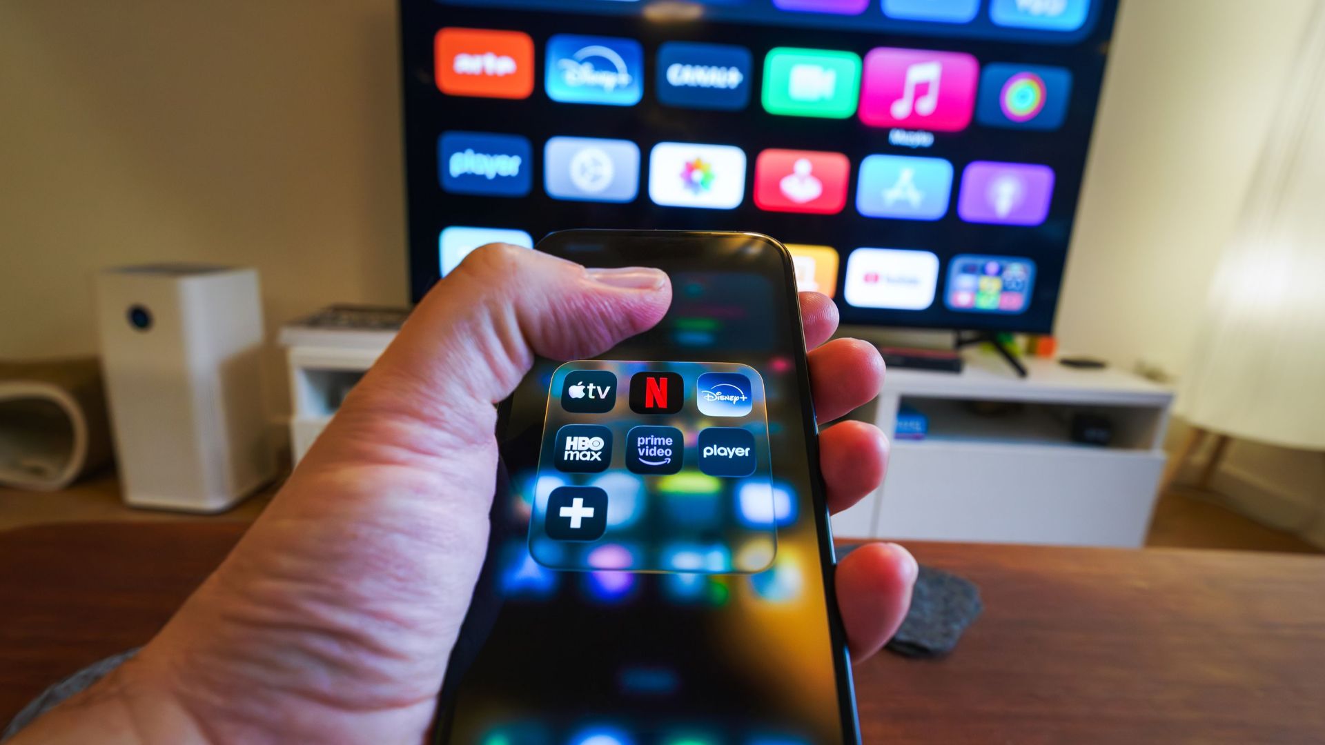 Close-up of a hand holding a phone displaying streaming apps in front of a TV with multiple app icons.