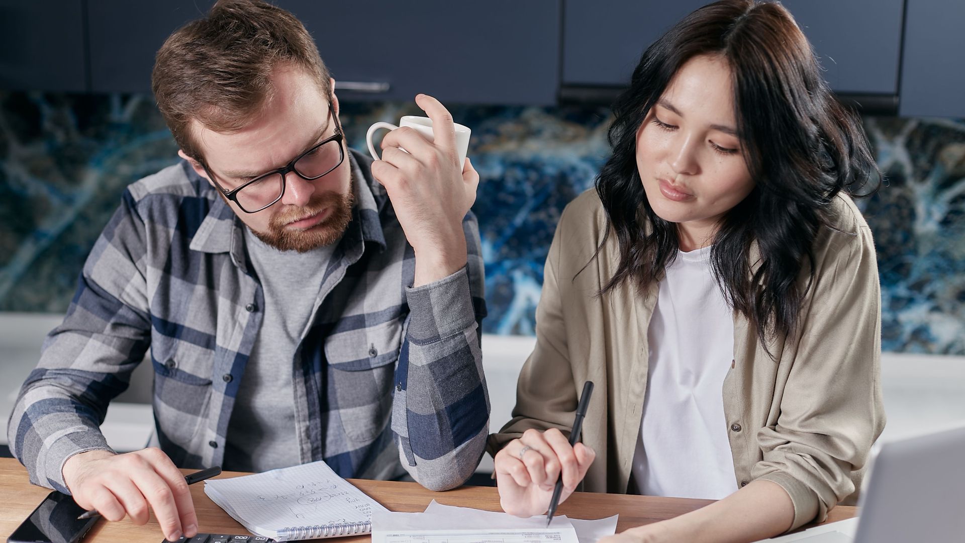 A couple reviewing household bills and budget using a calculator and laptop at their kitchen table.