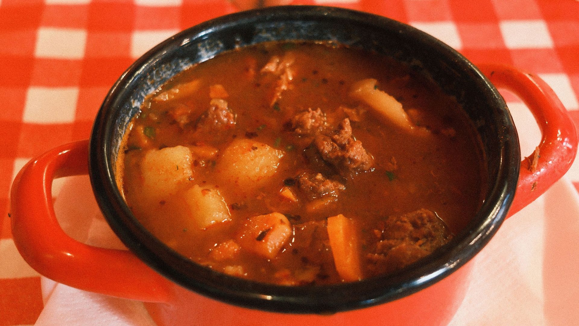A warm dish of Hungarian goulash in a red pot on a checkered tablecloth.