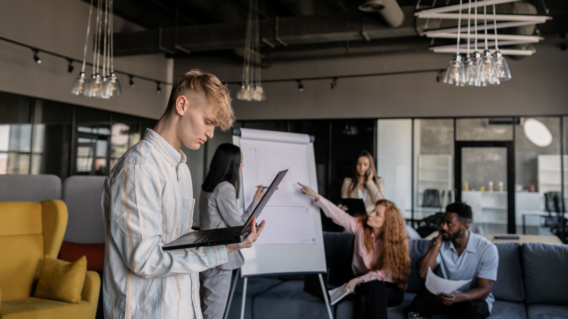 Side view of concentrated young male employee using laptop near group of diverse colleagues discussing project during brainstorm session in creative workplace