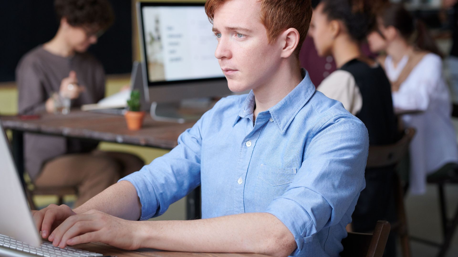 Young professional working intently on a computer in a busy modern office setting, surrounded by colleagues.
