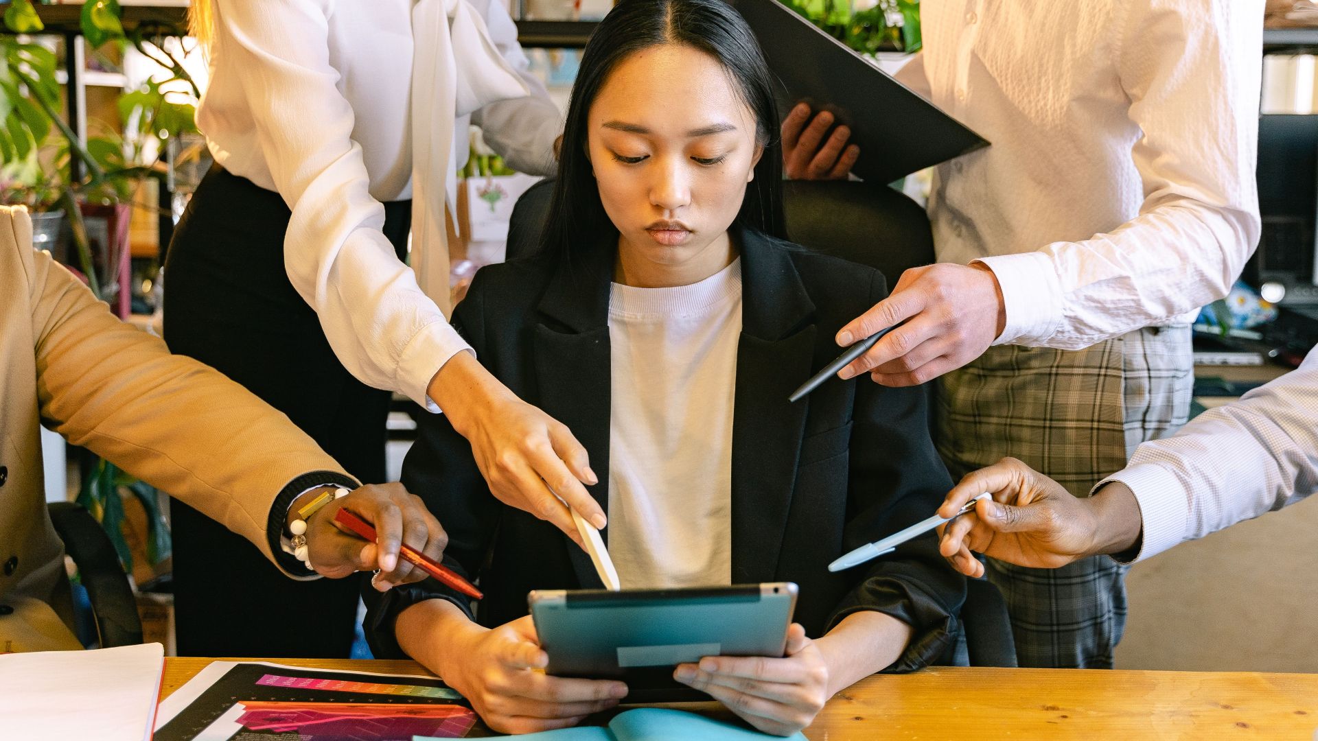Young woman working on a tablet, surrounded by colleagues seeking attention in a busy office setting.