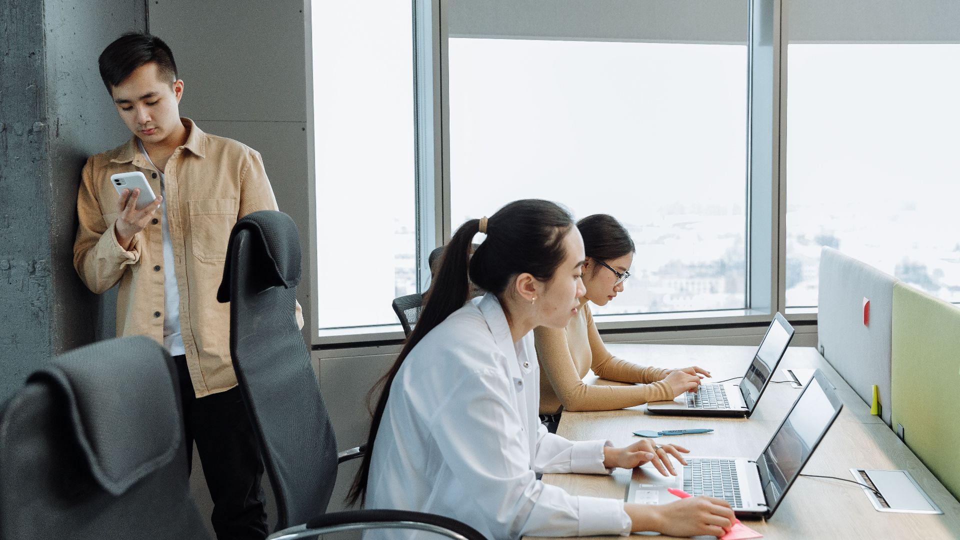 Three professionals working on laptops in a modern office setting with large windows.