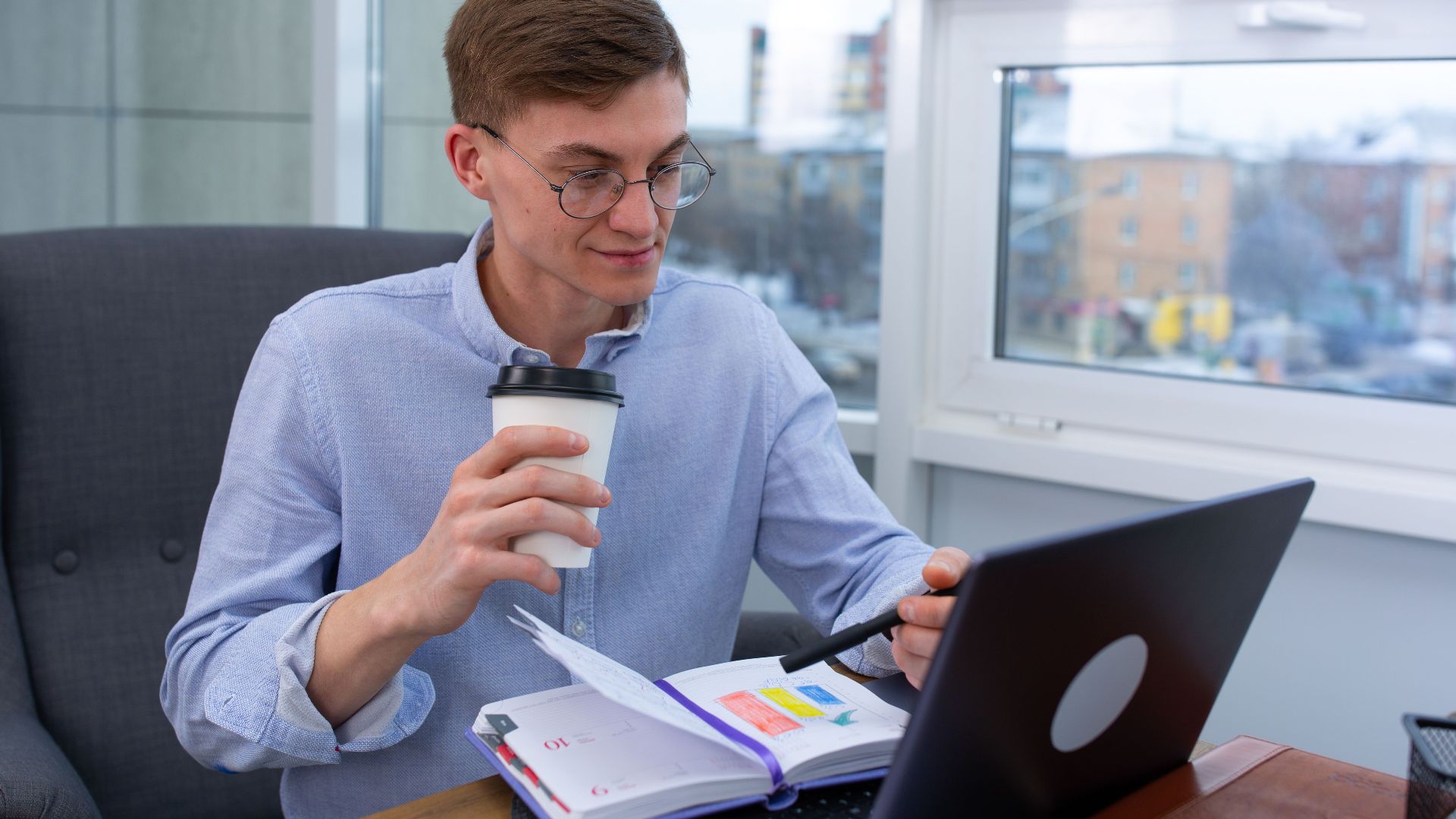 Business professional sipping coffee while working on a laptop with a planner at an office.