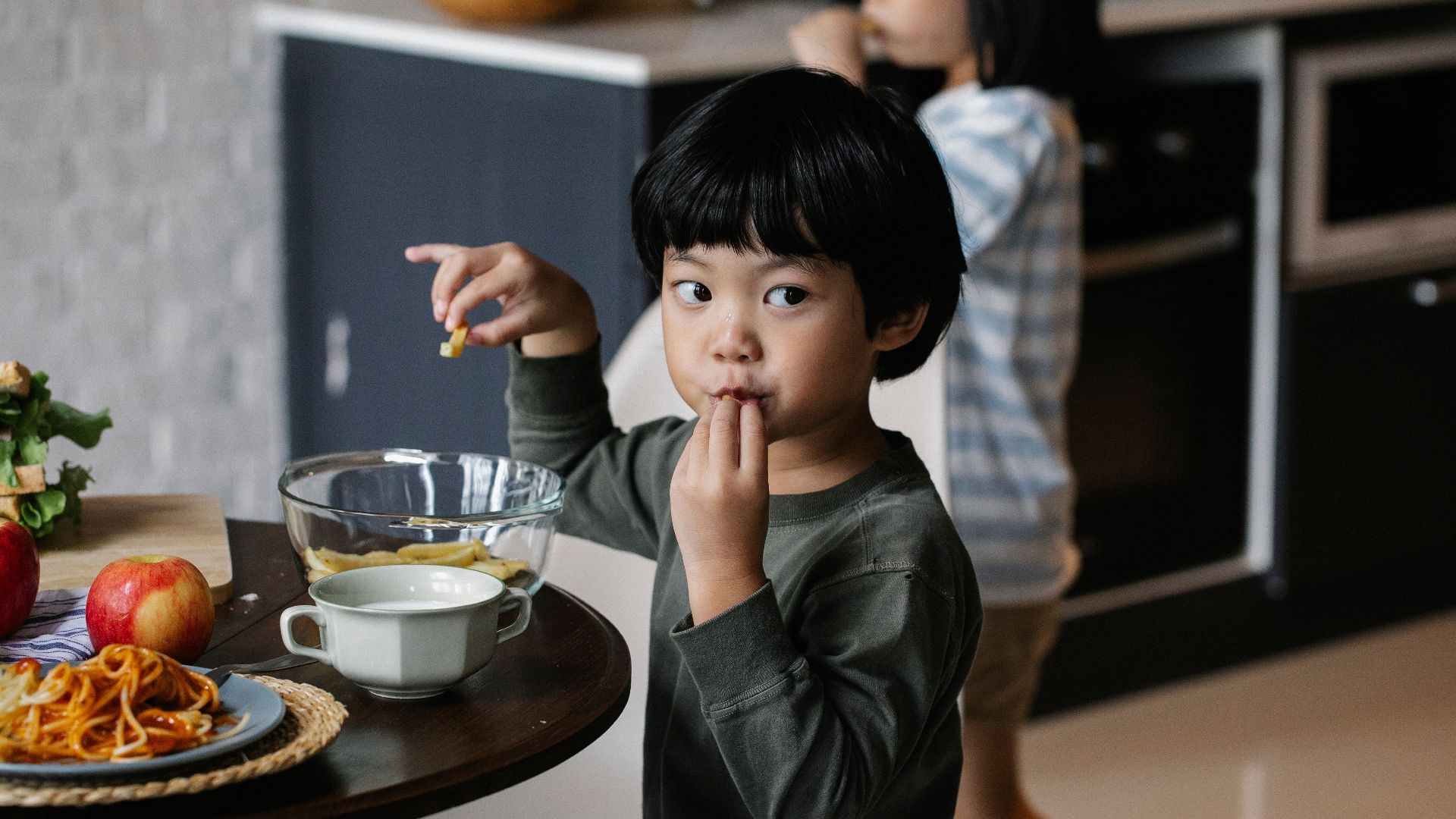 Little boy in casual clothes standing near table in kitchen while eating delicious homemade food and looking away
