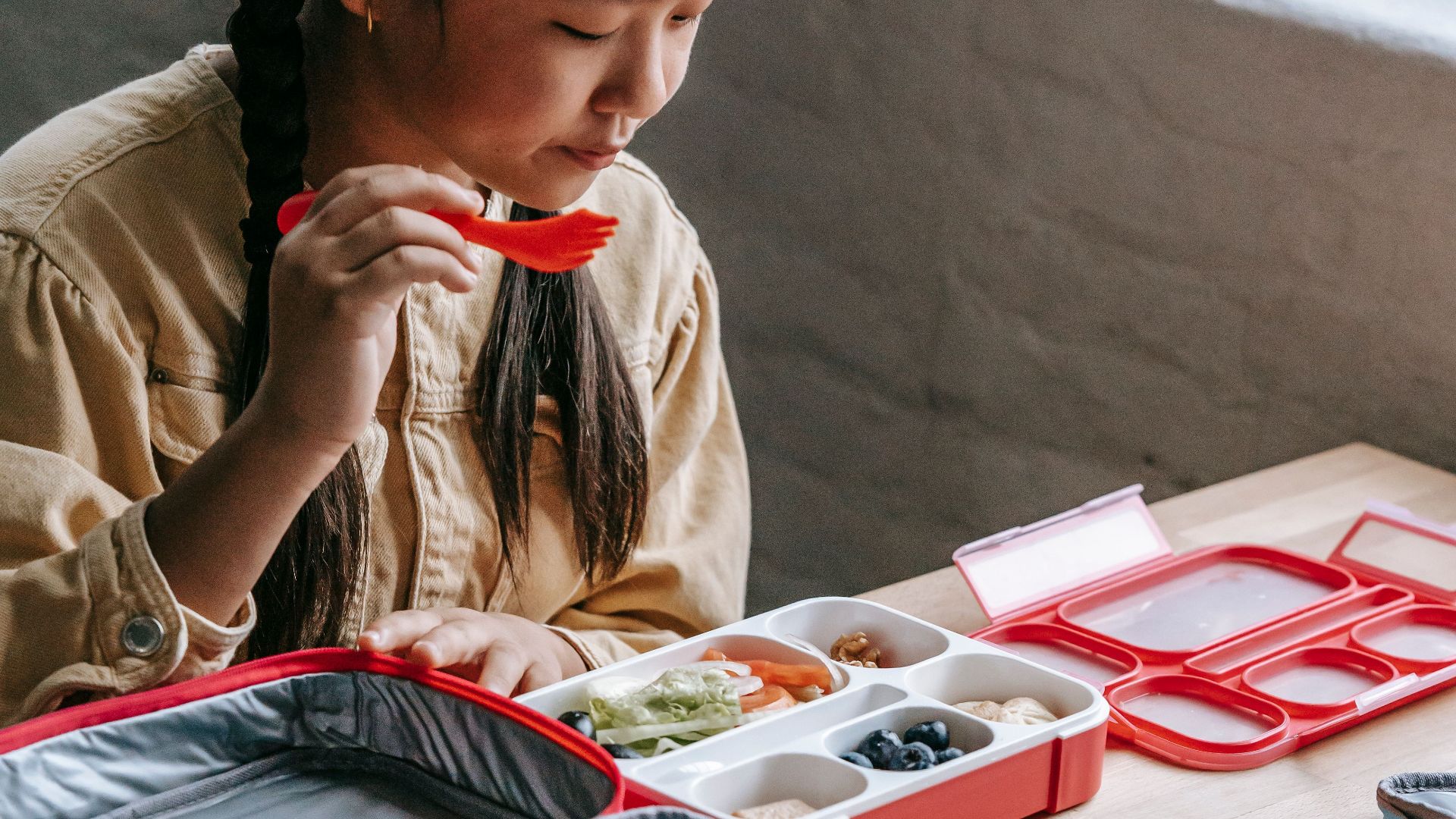 Crop adorable ethnic girl choosing healthy food from plastic container while sitting at table