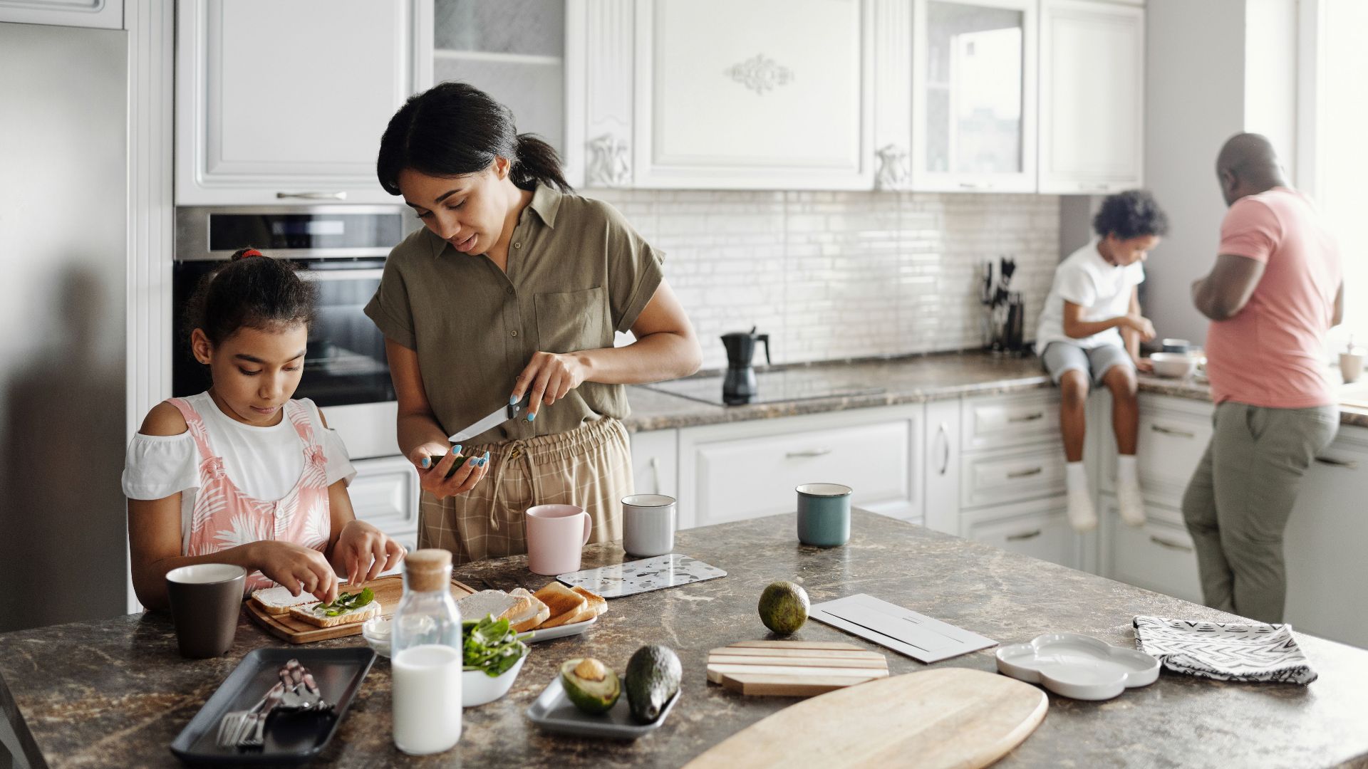 A mother and daughter preparing avocado toast in a bright kitchen while the father and son interact in the background.