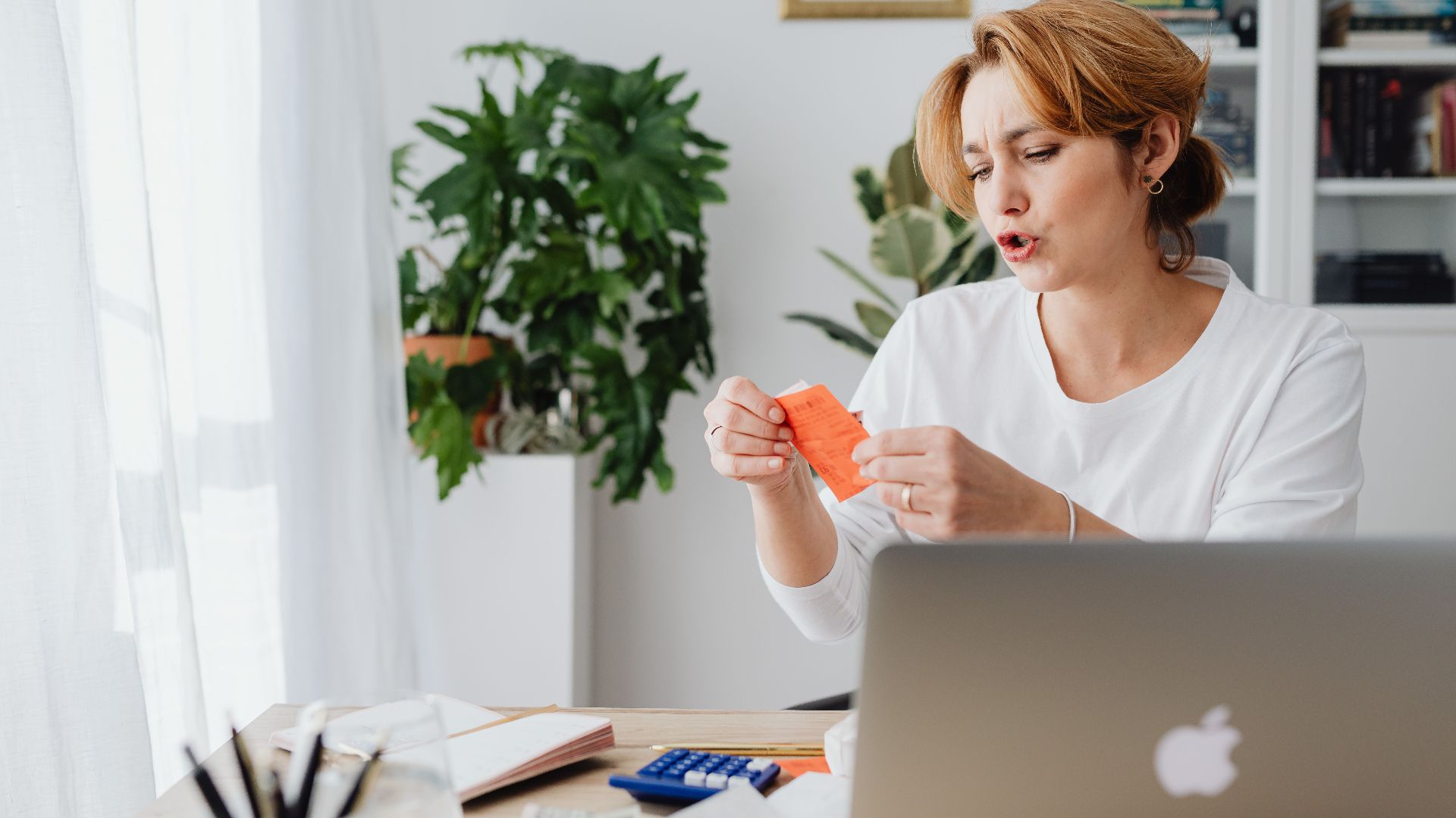 Woman reviewing bills at home desk with laptop and plants, managing personal finances.