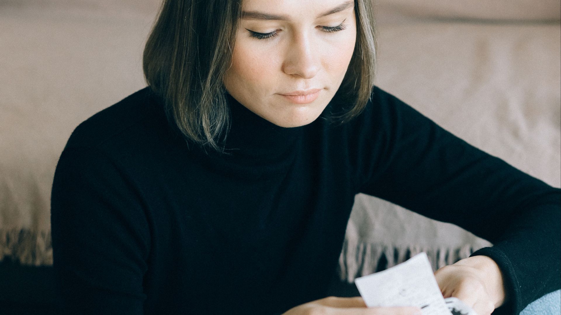 Young woman in black sweater reviewing bills at home, using a calculator, surrounded by documents.
